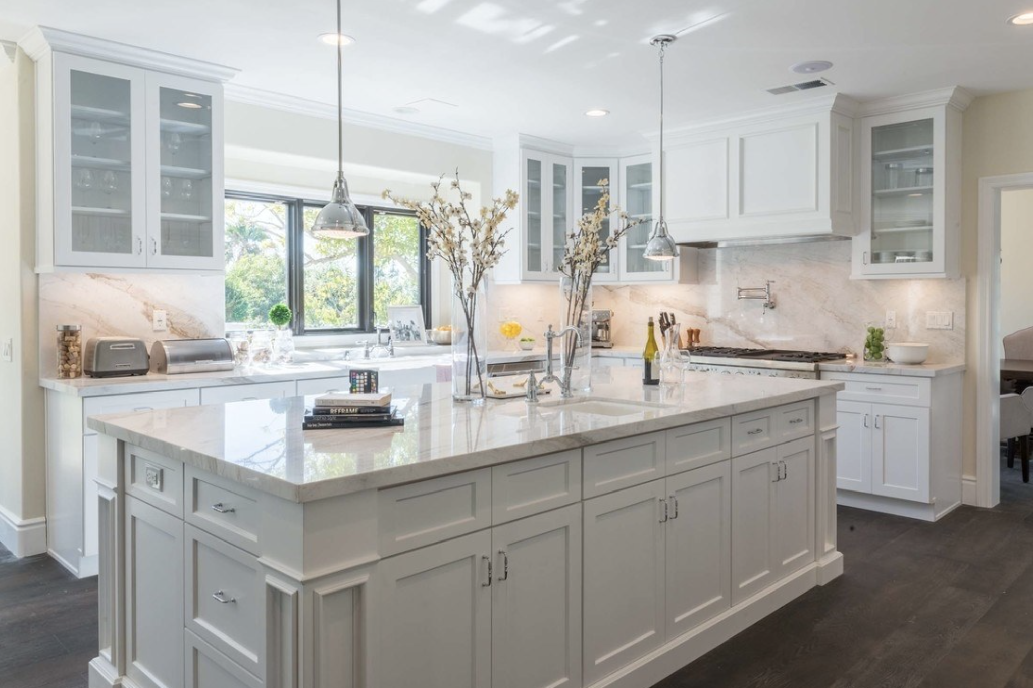 Cocina moderna con isla central, gabinetes blancos, repisa de ventanas con vista a árboles, iluminación empotrada, y decoración con flores y utensilios de cocina.