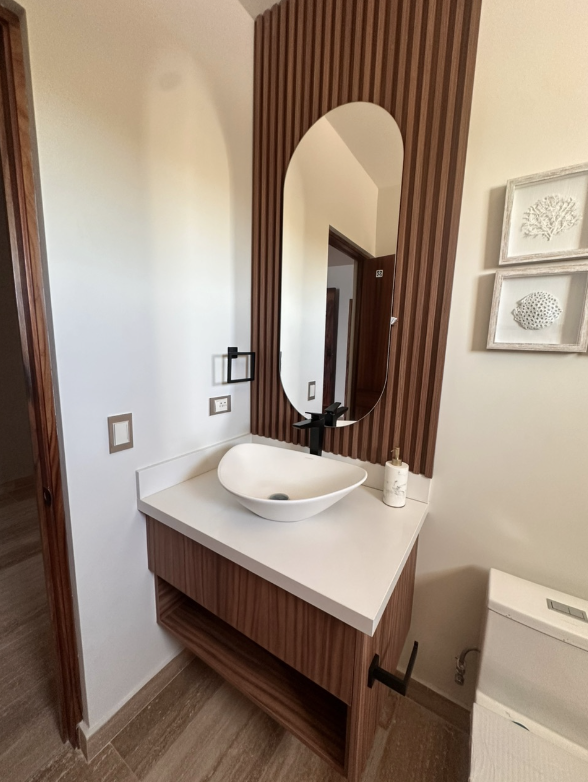 Lavabo sobre una encimera blanca con espejo ovalado y decoración de madera en la pared y cuadros con motivos marinos.