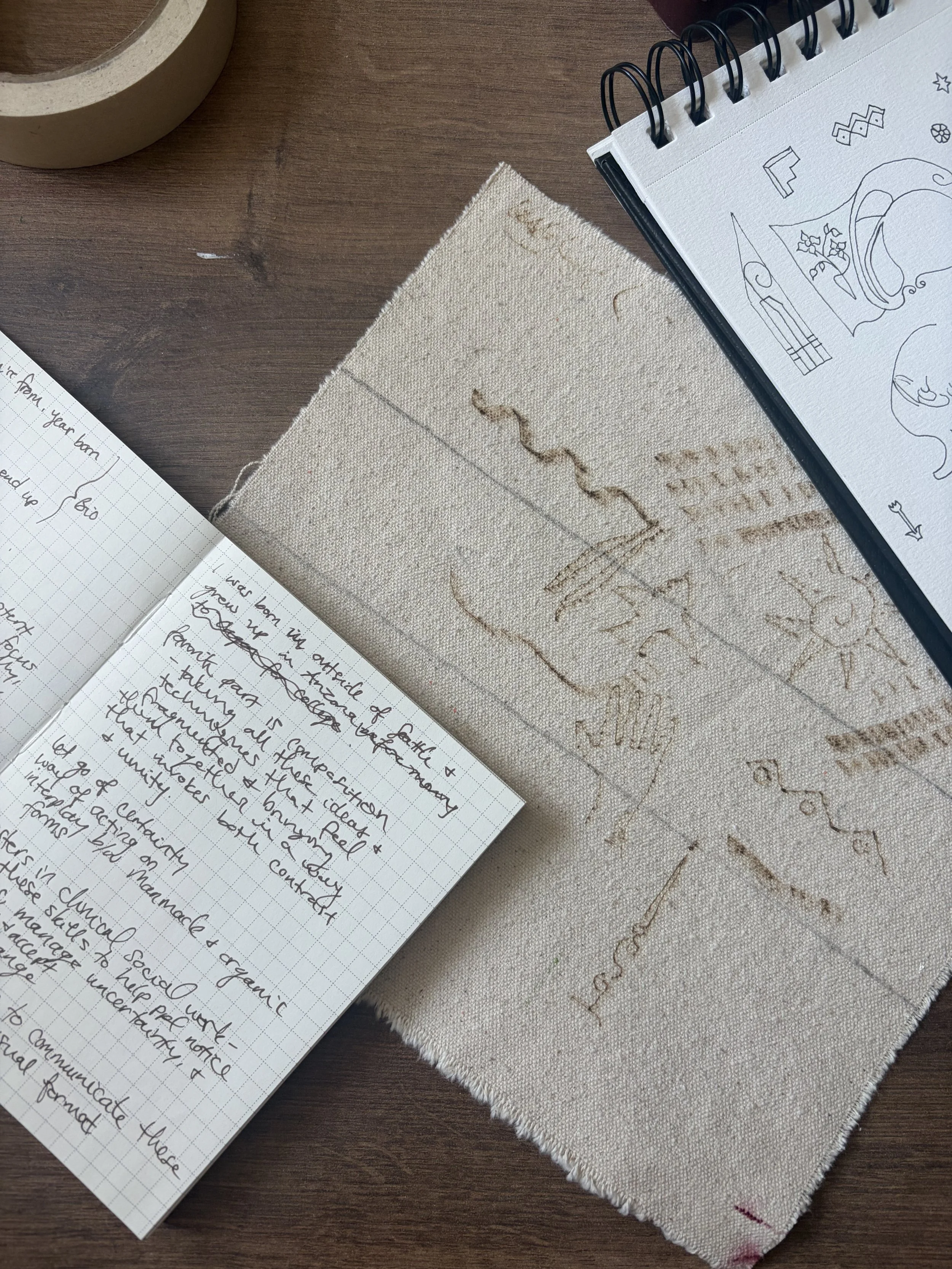 A close-up photograph of handwritten notes on grid paper, a piece of embroidered fabric, and a spiral-bound sketchbook with doodles, all on a wooden surface.