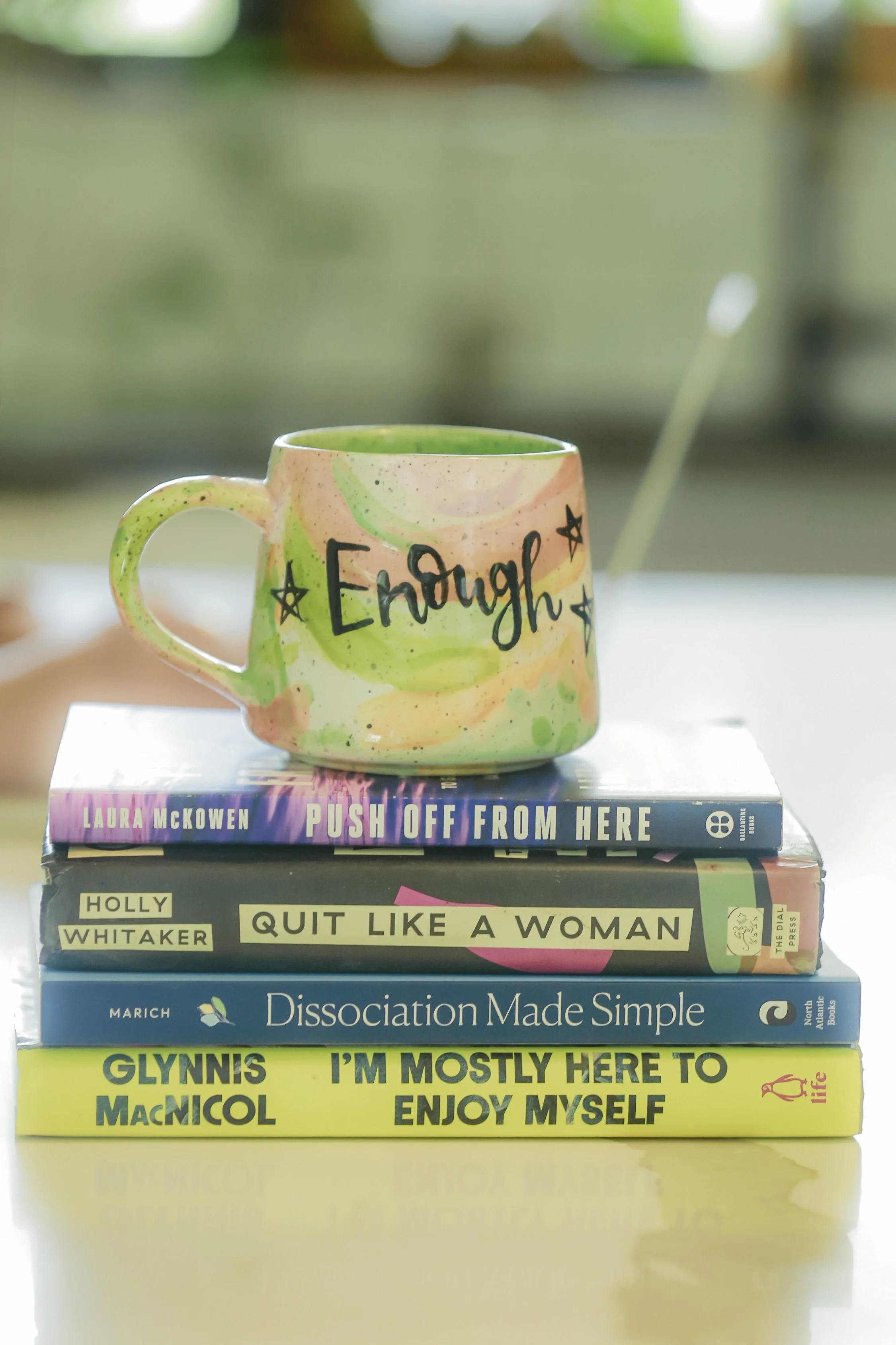 Stack of five books with a colorful mug on top. The mug has the word 'Enough' written on it. The books are titled 'Push Off From Here' by Laura McKowen, 'Quit Like a Woman' by Holly Whitaker, 'Dissociation Made Simple' by Marich, and 'Glynis I'm Mostly Here to Enjoy Myself' by Glynis McNicol.