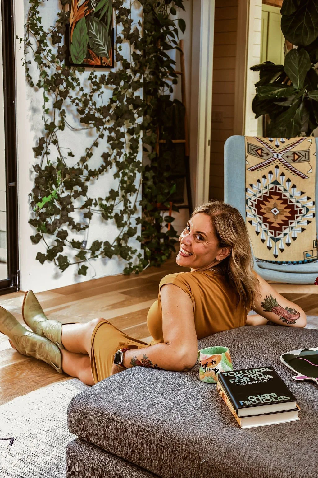 A woman in a mustard-colored dress and knee-high boots is lying on her stomach on a gray ottoman in a cozy, decorated room. She has tattoos on her arms and is smiling at the camera. There are houseplants, a patterned chair with a southwestern design, and books on the ottoman in the background.