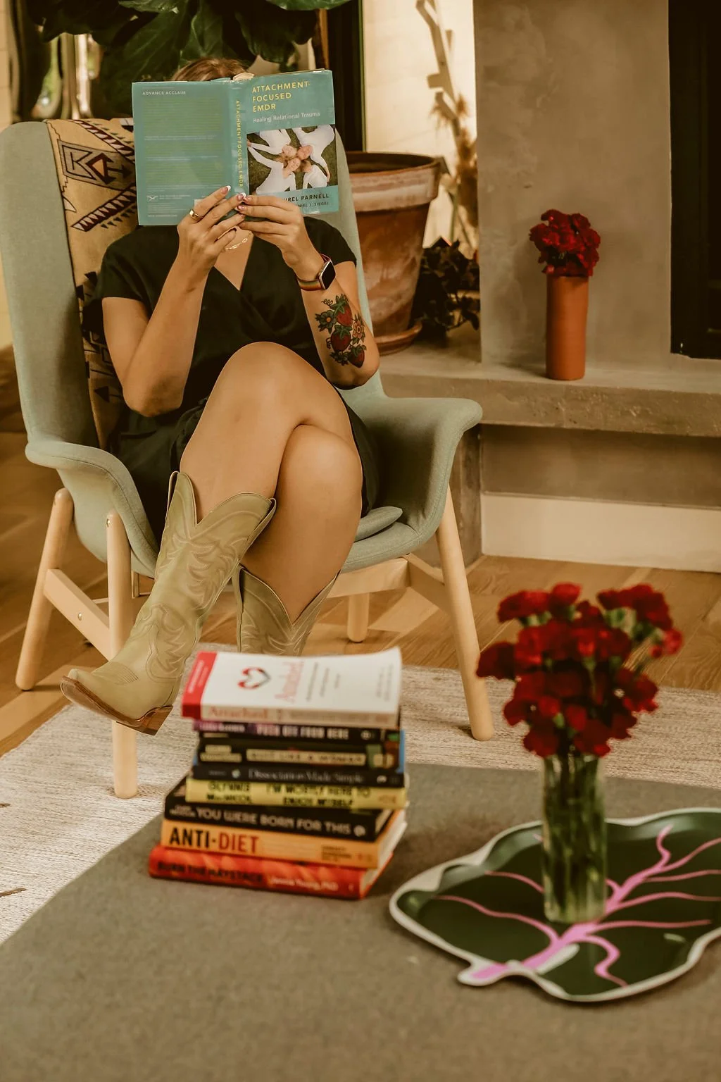 Woman sitting in a modern living room chair reading a teal book, wearing cowboy boots, a black dress, a smartwatch, and displaying a tattoo on her arm. There is a stack of books and a vase with red flowers on a table in front of her.