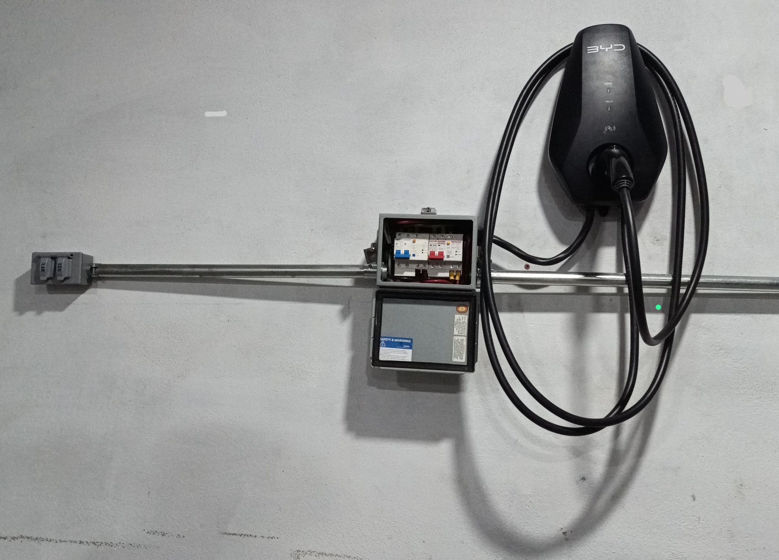 Instalación eléctrica en una pared con un enchufe, un regulador y una caja de conexiones con cables conectados, además de una caja de disyuntores con interruptores de diferentes colores.