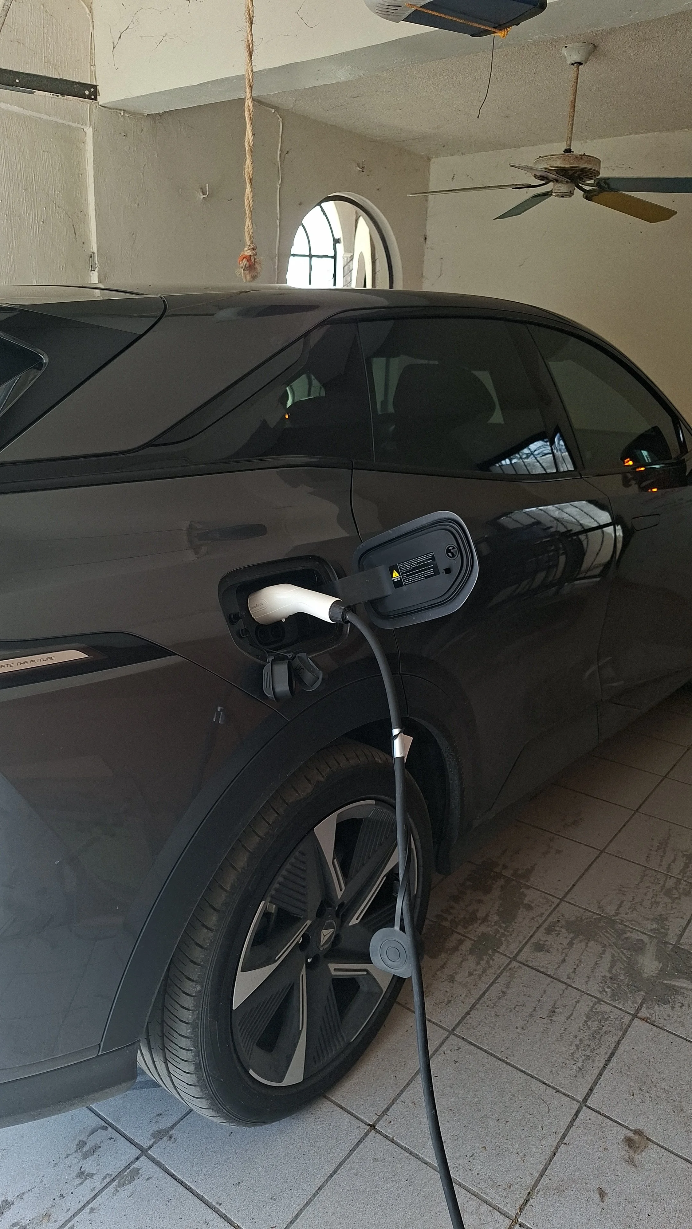Auto negro enchufado a cargador de vehículo eléctrico en un garaje con piso de baldosas y ventiladores en el techo.