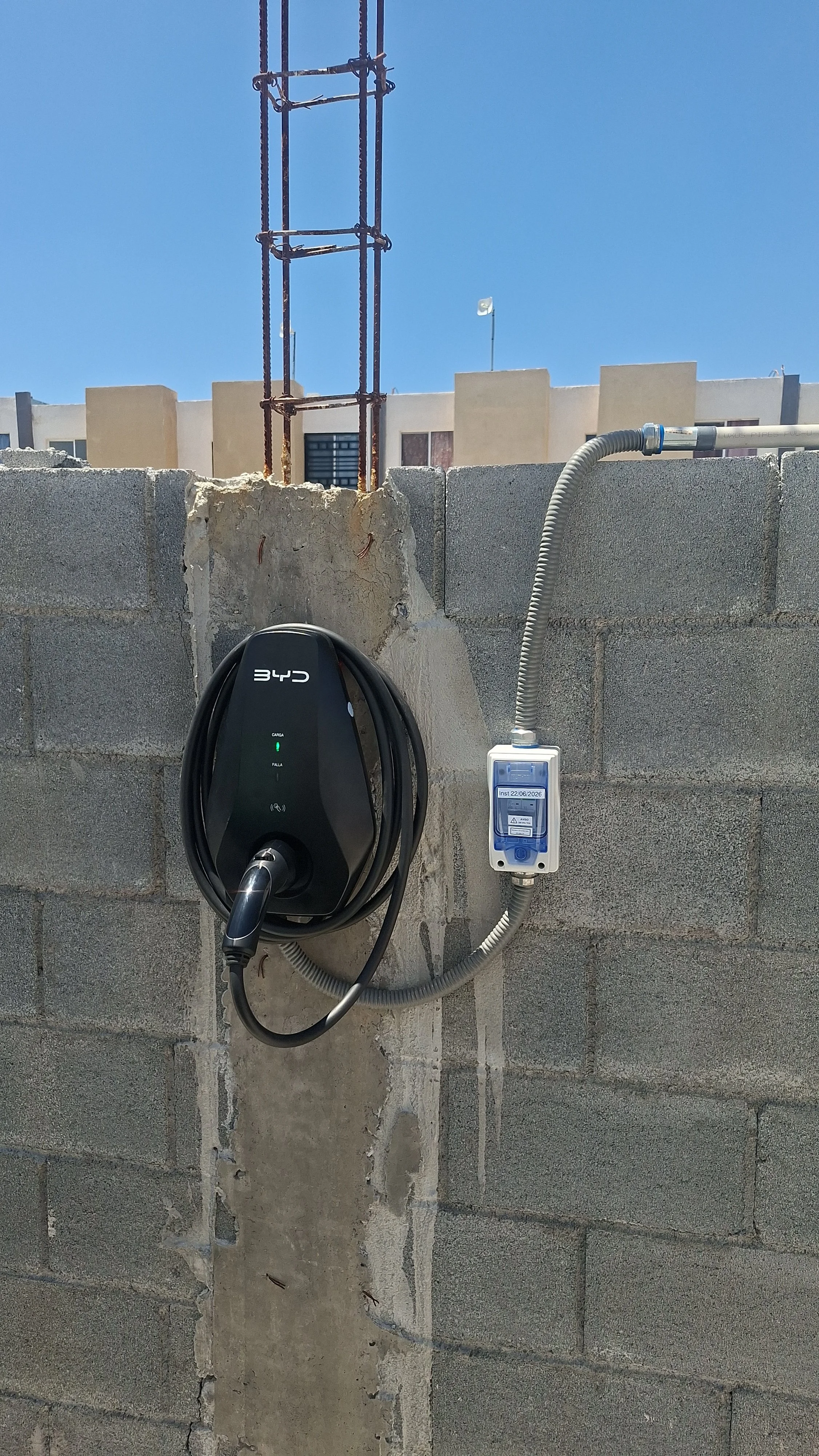 Cochera con pared de bloques, en la pared hay un cargador de coche eléctrico y una toma de corriente con cable conectado, en el fondo se ve un muro de construcción y un cielo despejado.