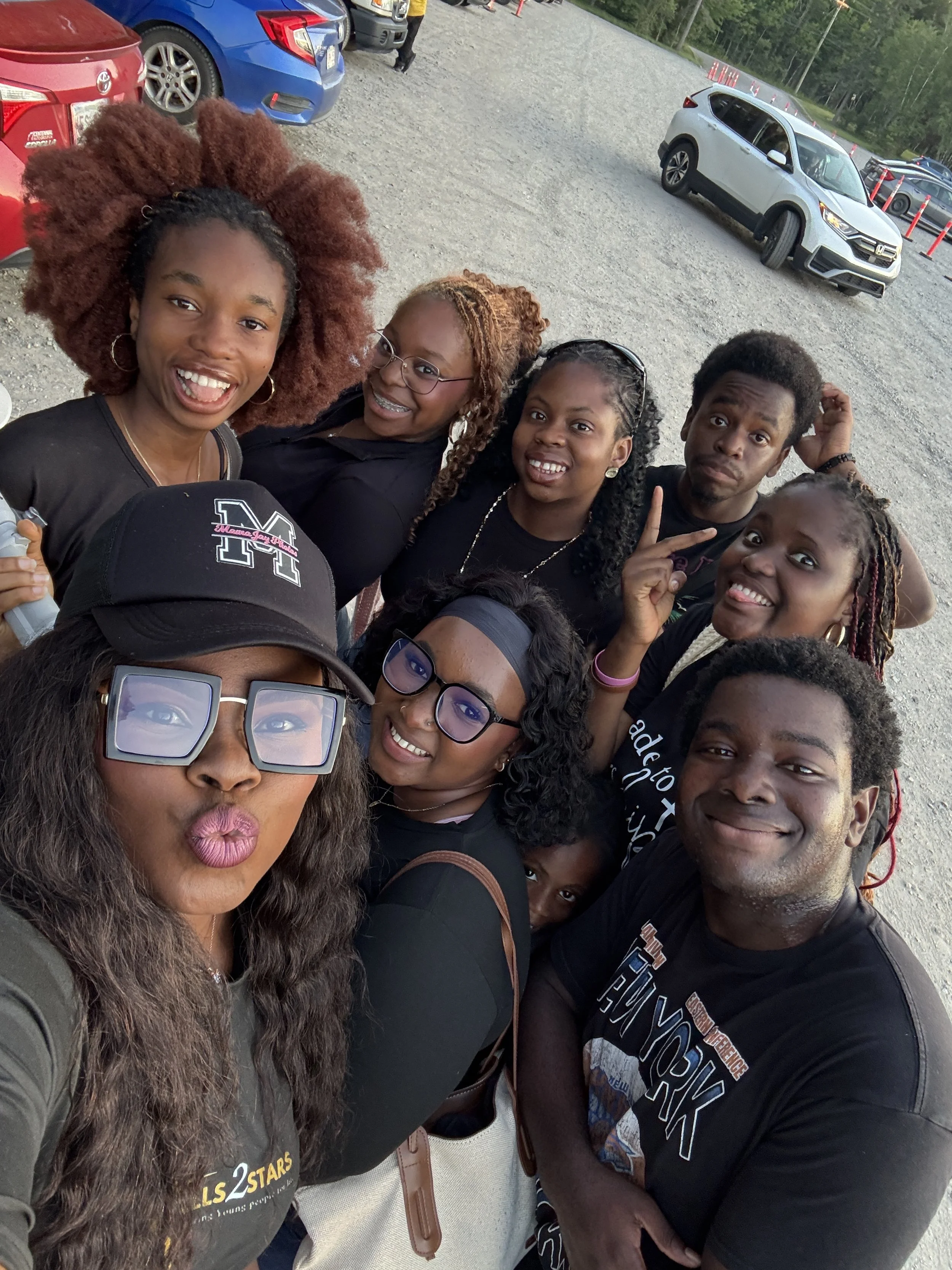 Group of smiling people taking a selfie in a parking lot with several cars and trees in the background.