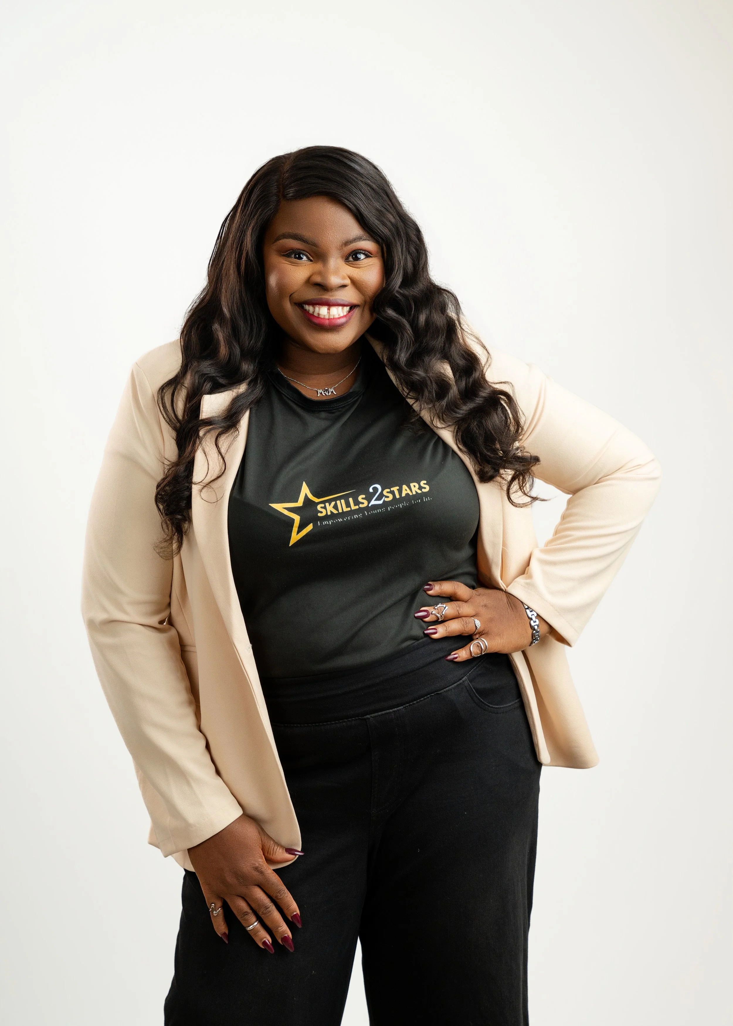 A woman with long curly black hair wearing a black t-shirt with a yellow star and the text "Skills 2 Stars" and a beige blazer, standing against a plain white background, smiling, with her right hand on her hip and her left hand resting on her thigh.