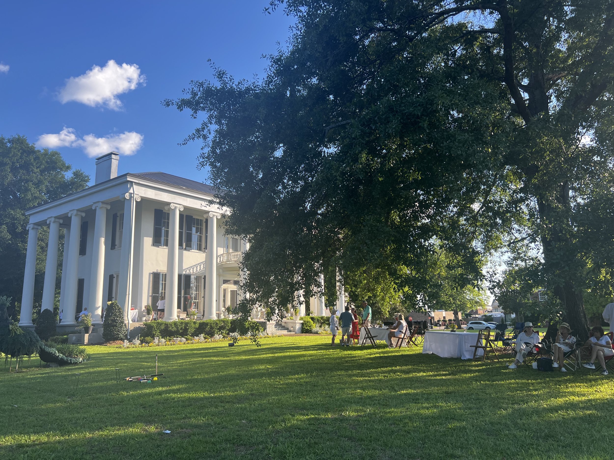 White historic mansion with tall columns, green lawn, and people gathered outdoors under trees on sunny day.