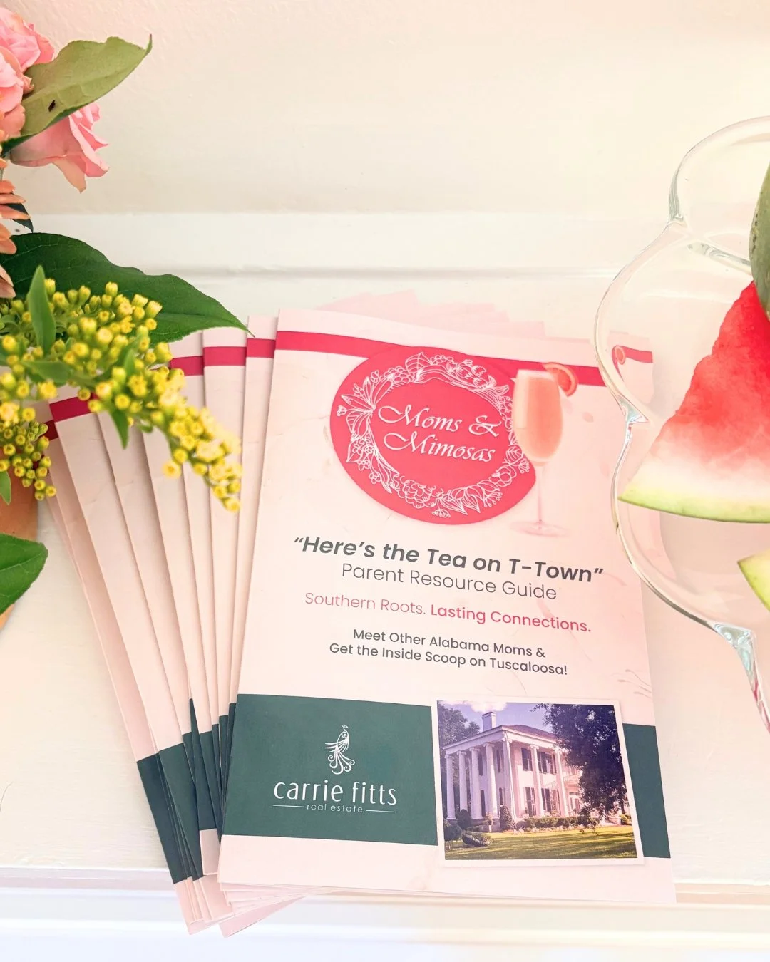 Pamphlets titled 'Here's the Tea on T-Town' Parent Resource Guide with a pink circular logo 'Moms & Mimosas' and a champagne glass illustration, surrounded by flowers and a glass bowl with watermelon slices.