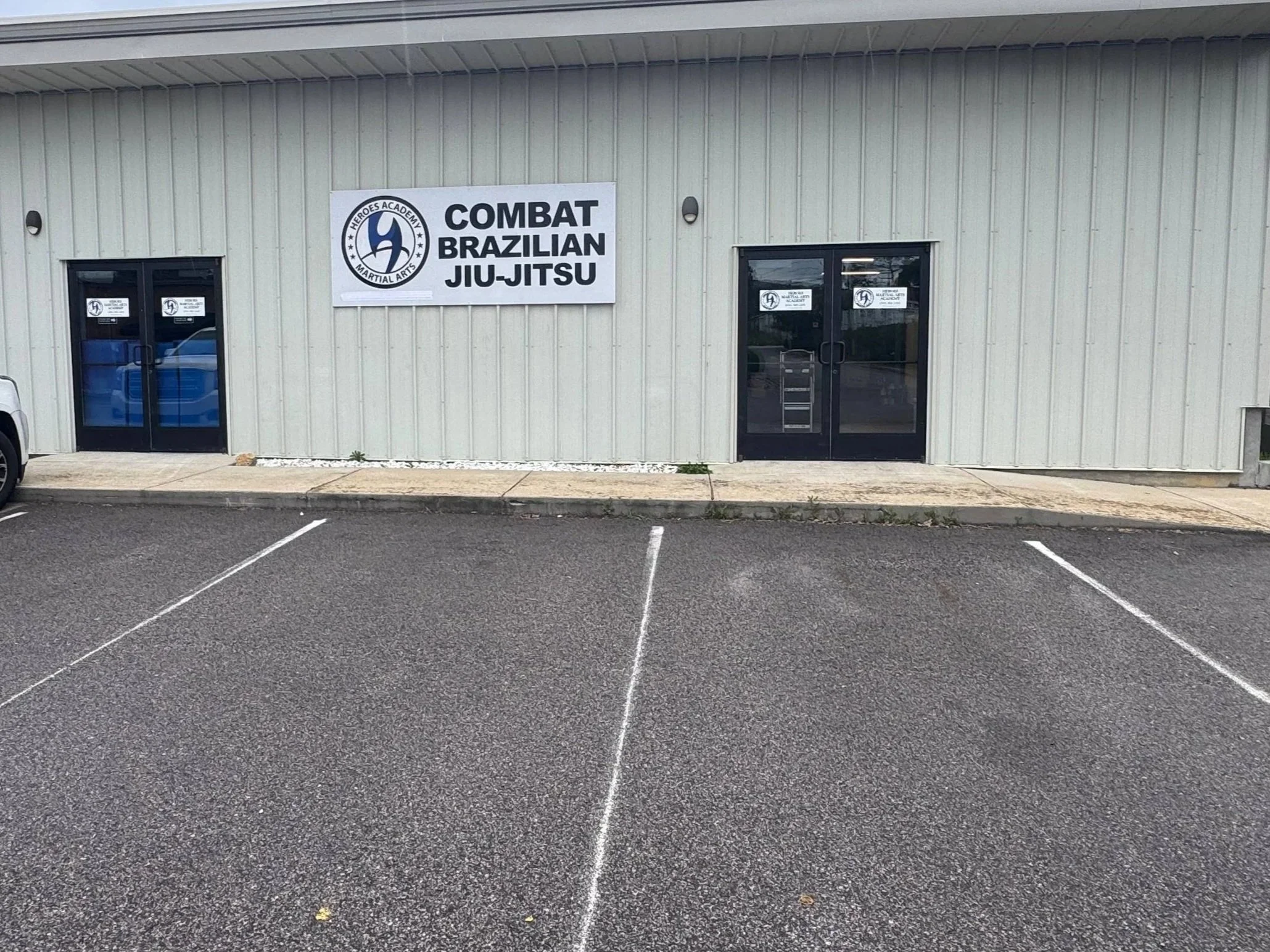 Exterior of Heroes Academy martial arts school with a large sign reading 'Heroes Academy Combat Brazilian Jiu-Jitsu' and two glass doors, parking lot in front.