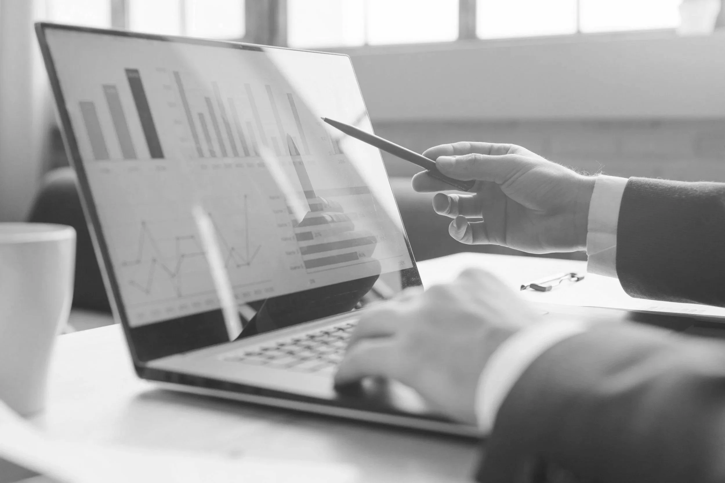 Black and white image of a person's hand pointing a stylus at a tablet displaying bar graphs and charts, on a desk with a laptop, a coffee mug, and a pair of glasses.