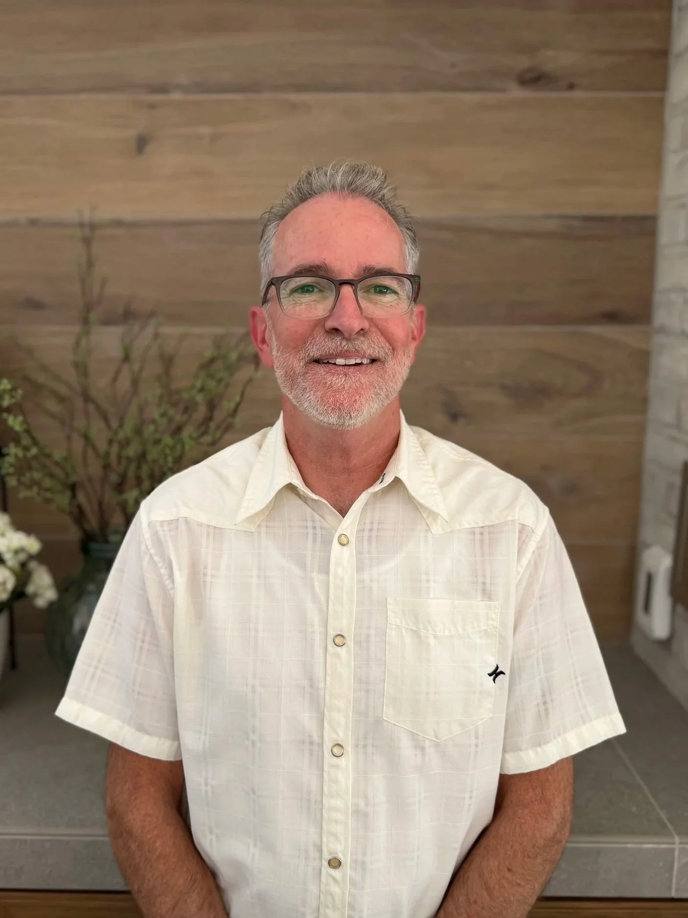 A man with brown and grey hair and beard, wearing glasses and a light-colored short-sleeve button-up shirt, standing indoors against a wooden panel wall with a plant in the background.