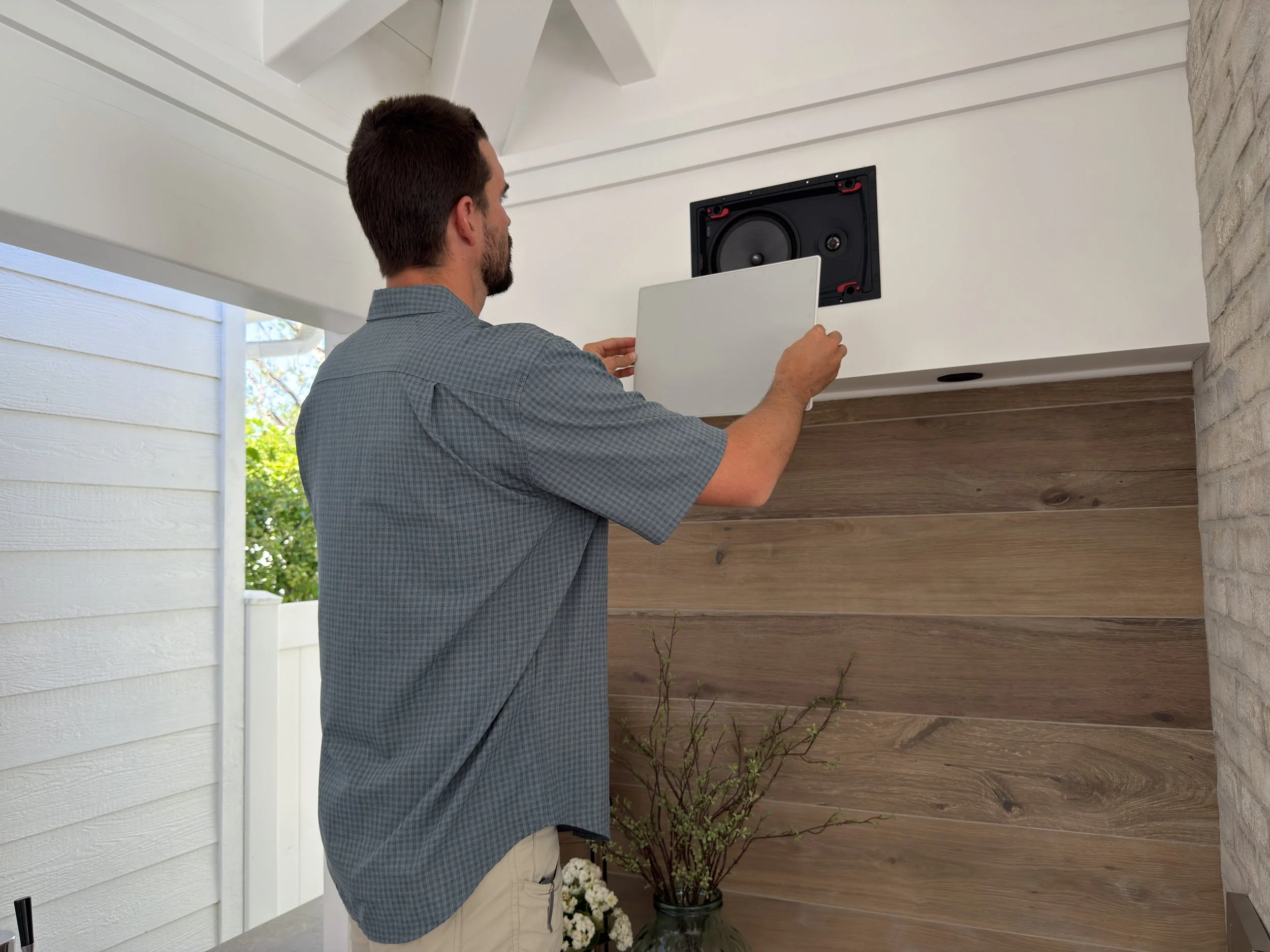 A man installing a white outdoor speaker in a backyard in Orange County , above a wooden panel, and a brick wall, with a plant with white flowers nearby.