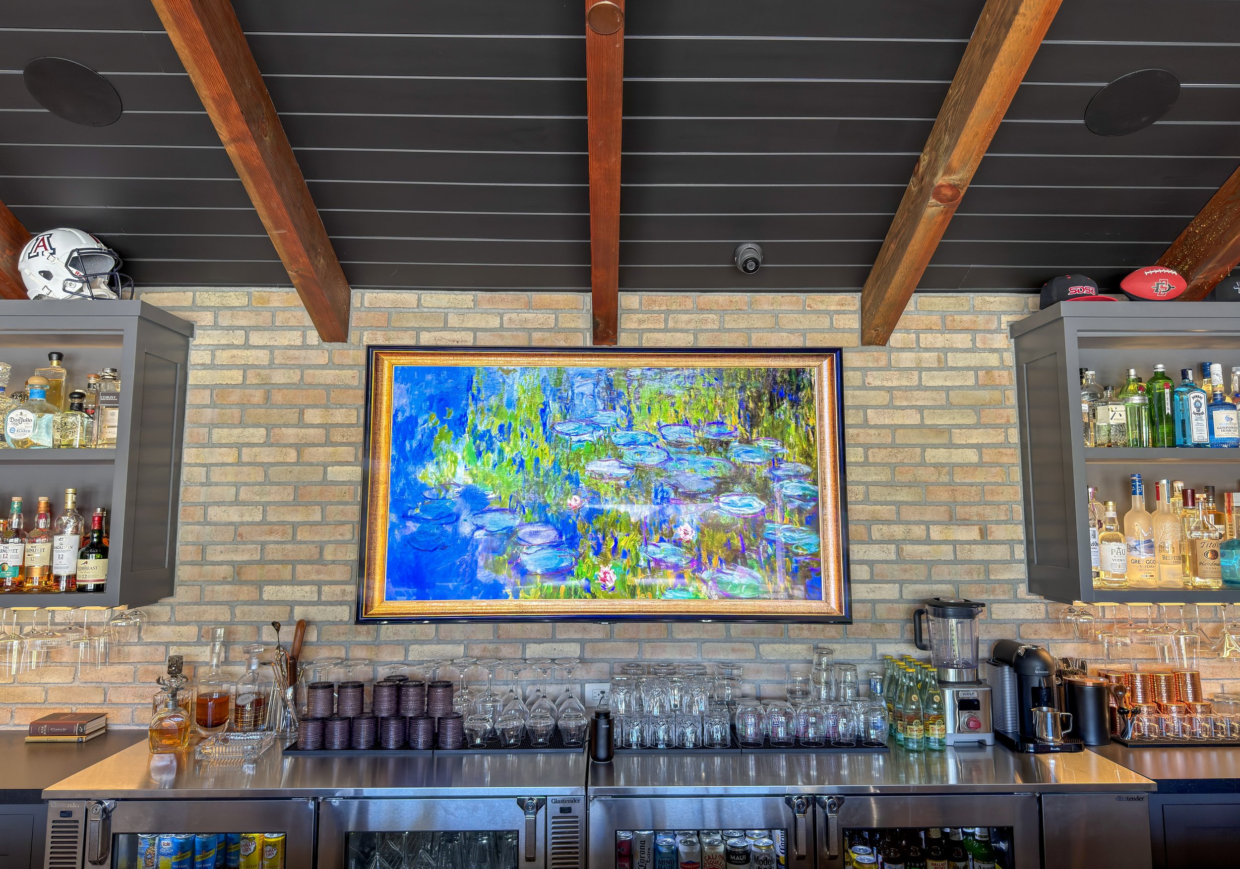 A bar area with liquor bottles on shelves, a colorful painting of a pond scene on a samsung tv, and a ceiling with exposed wooden beams.
