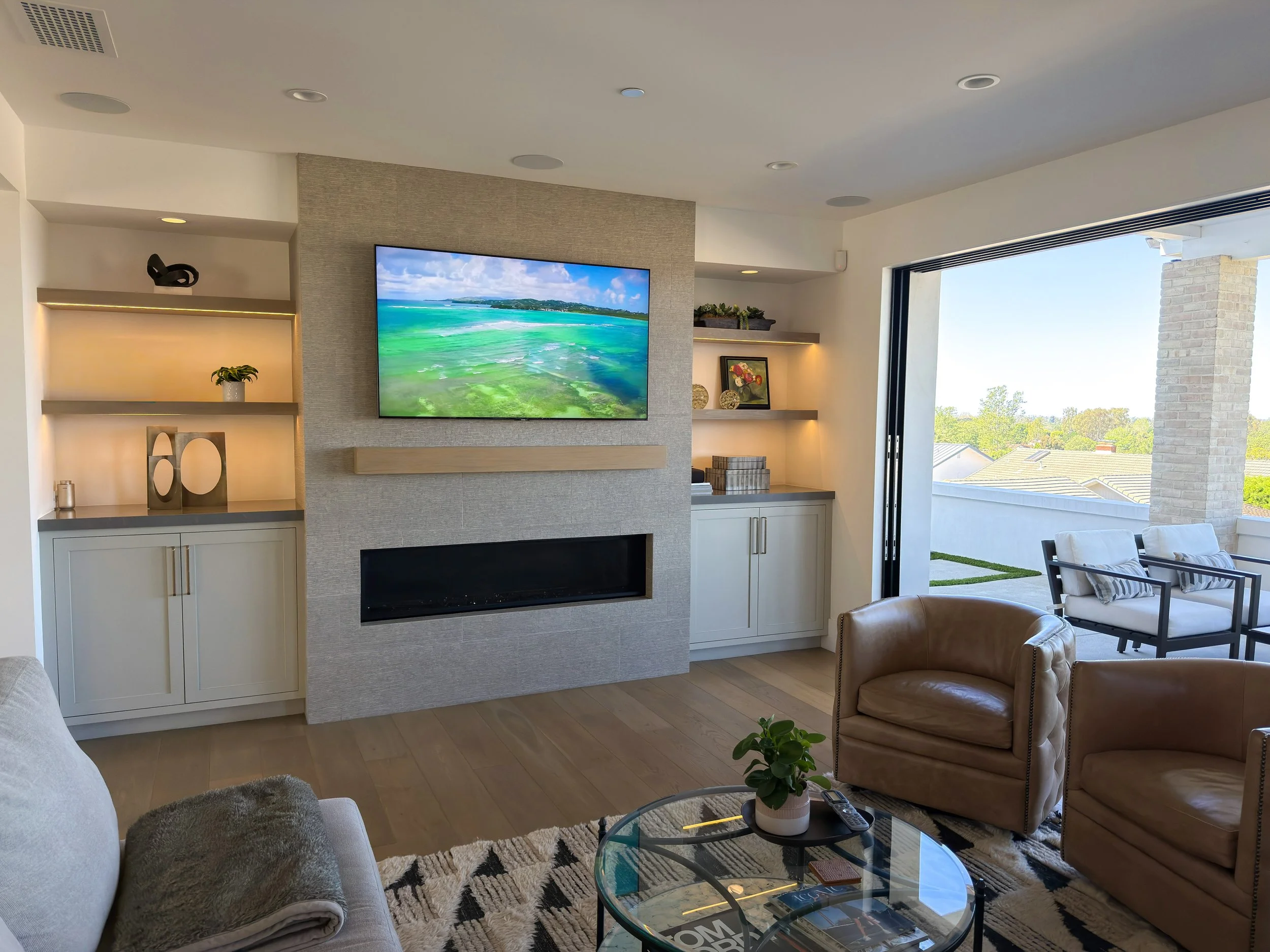 Living room with a built-in wall unit, a wall-mounted TV displaying a beach scene, beige armchairs, a glass coffee table, an open sliding door showing a patio with outdoor chairs and a view of trees and rooftops.
