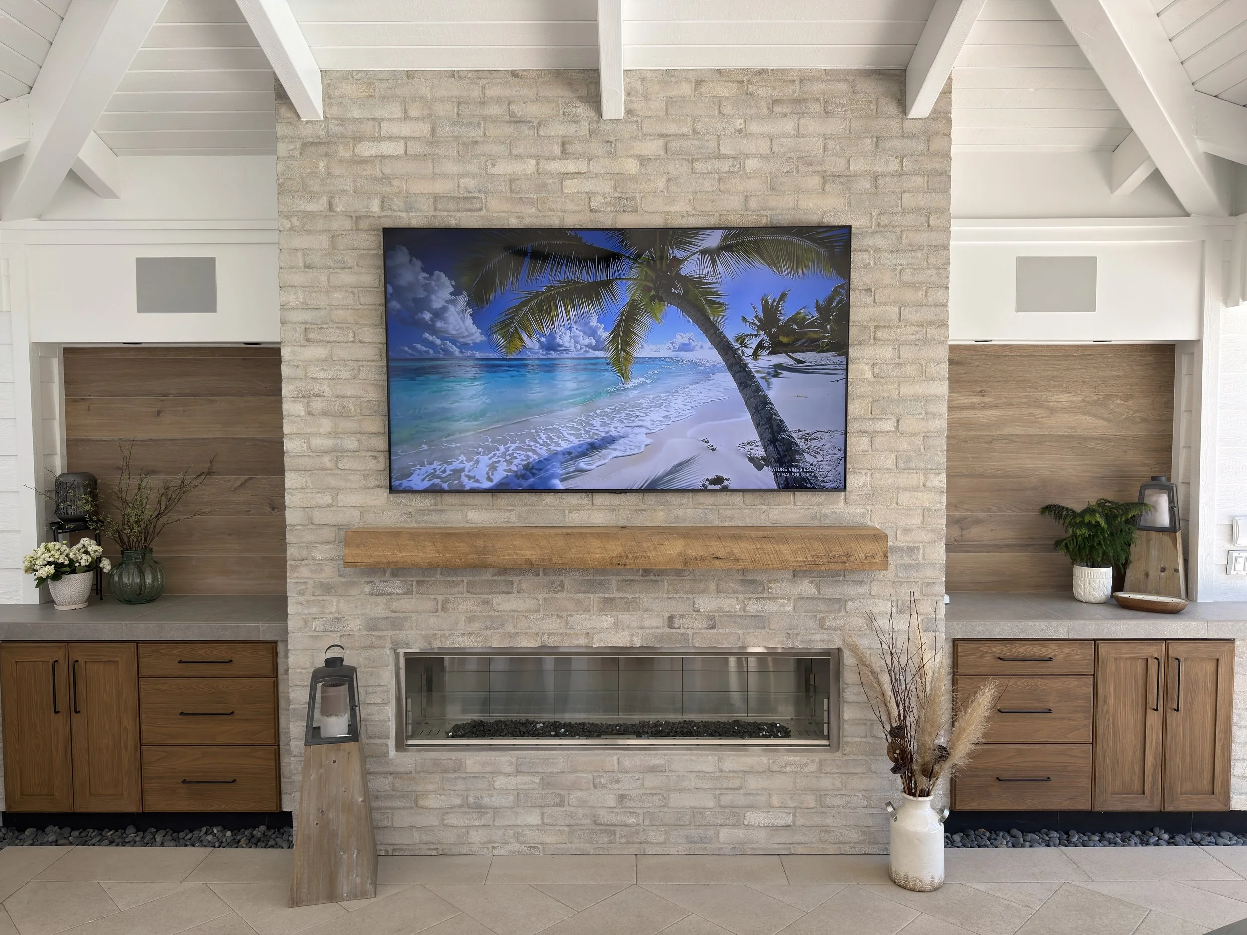 Living room with a large flat-screen TV showing a beach scene, set against a white brick fireplace, flanked by white and wooden cabinets, decorated with plants and vases.
