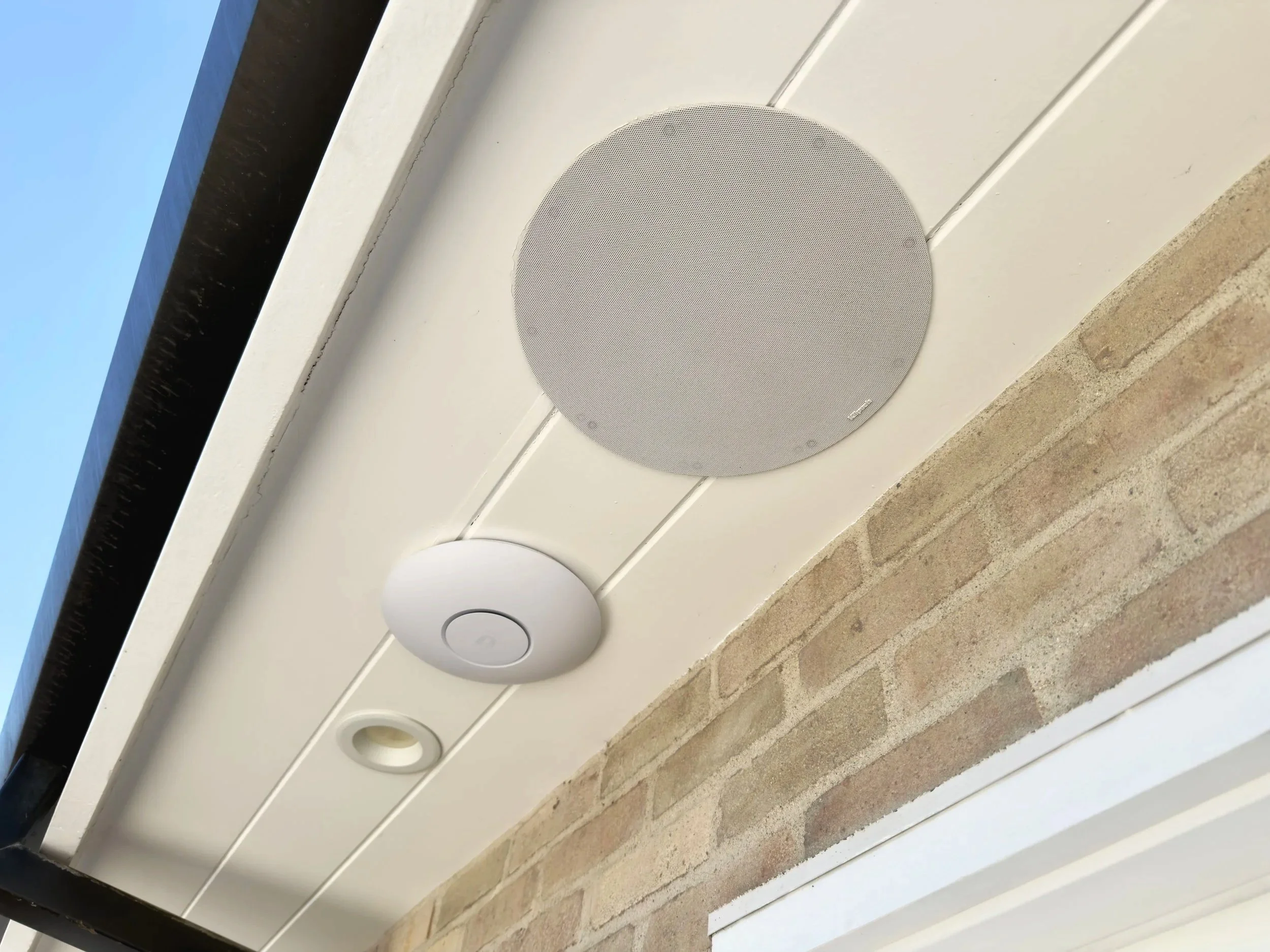 Close-up of a white outdoor wall with built-in circular speakers, lights, and wifi networking, with part of the sky and window frame visible in the background.