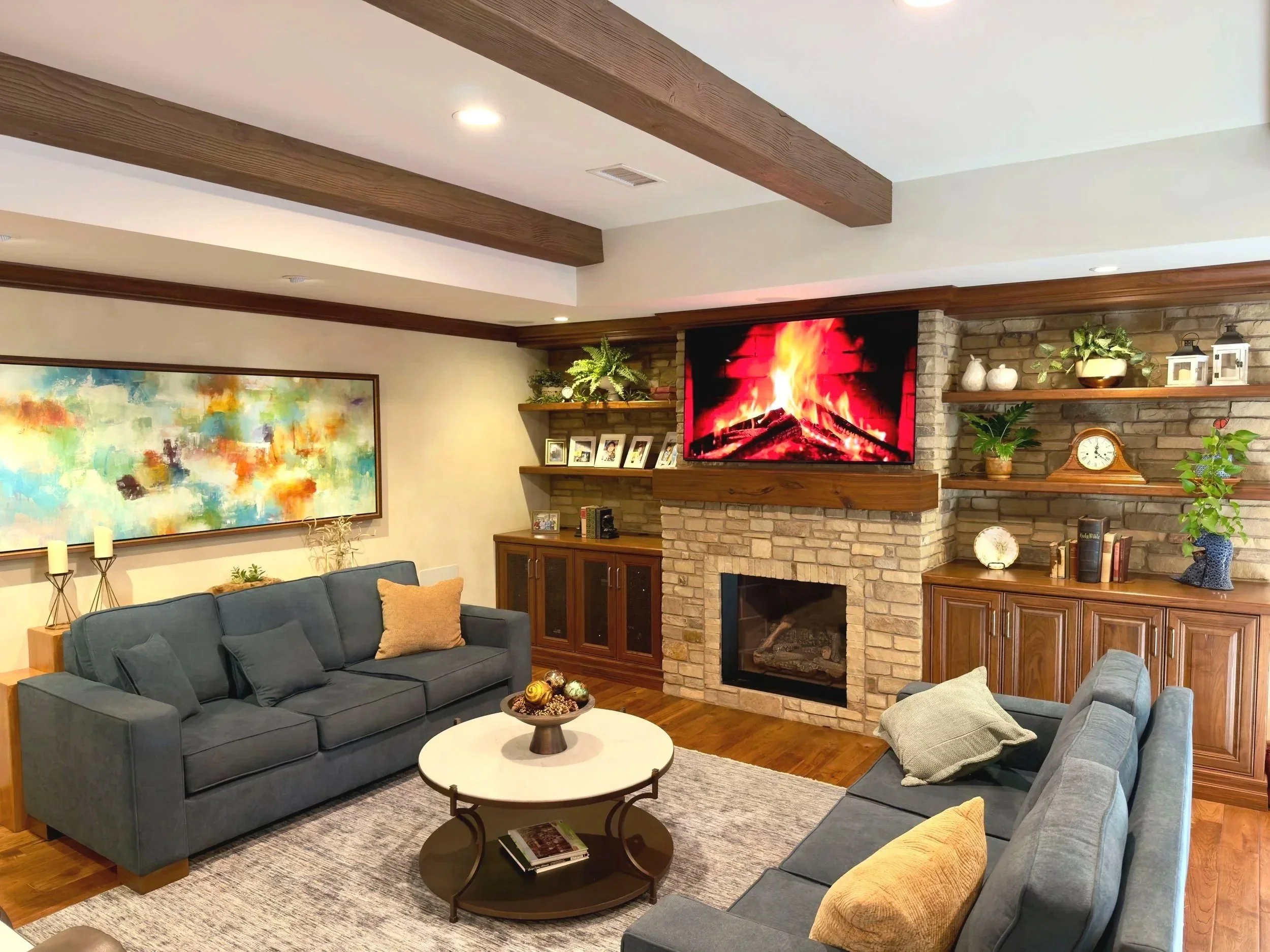 Living room with dark blue couch, patterned pillows, round coffee table with books and decorative bowl, fireplace with brick surround, wooden mantel with potted plants, picture frames, and decorative objects, colorful abstract painting on the wall, ceiling wooden beams, and a TV showing a fireplace scene.