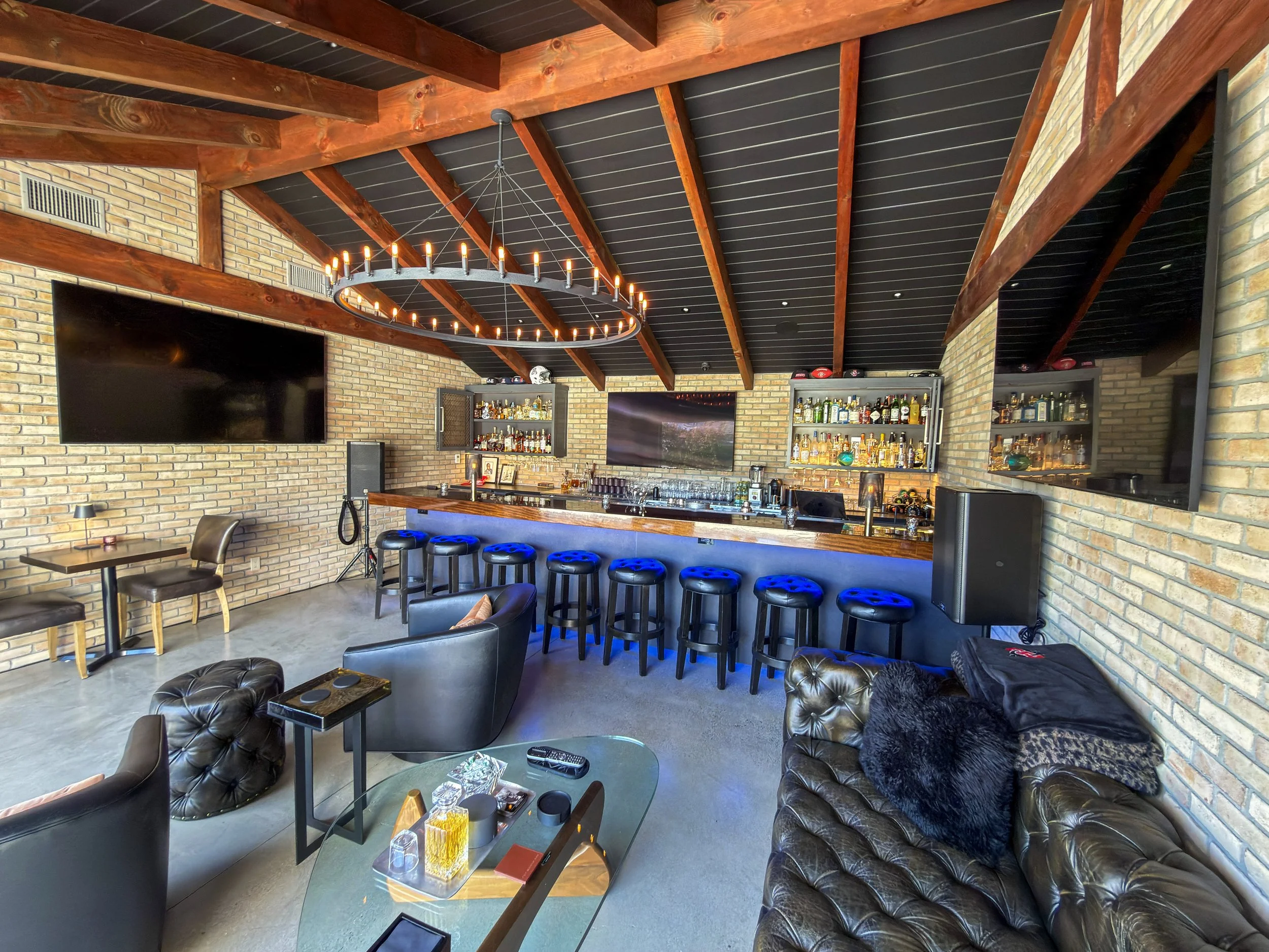 Custom home bar and entertainment lounge with mounted TVs, surround sound speakers, and exposed wood beam ceiling by Surf City Audio Visual.