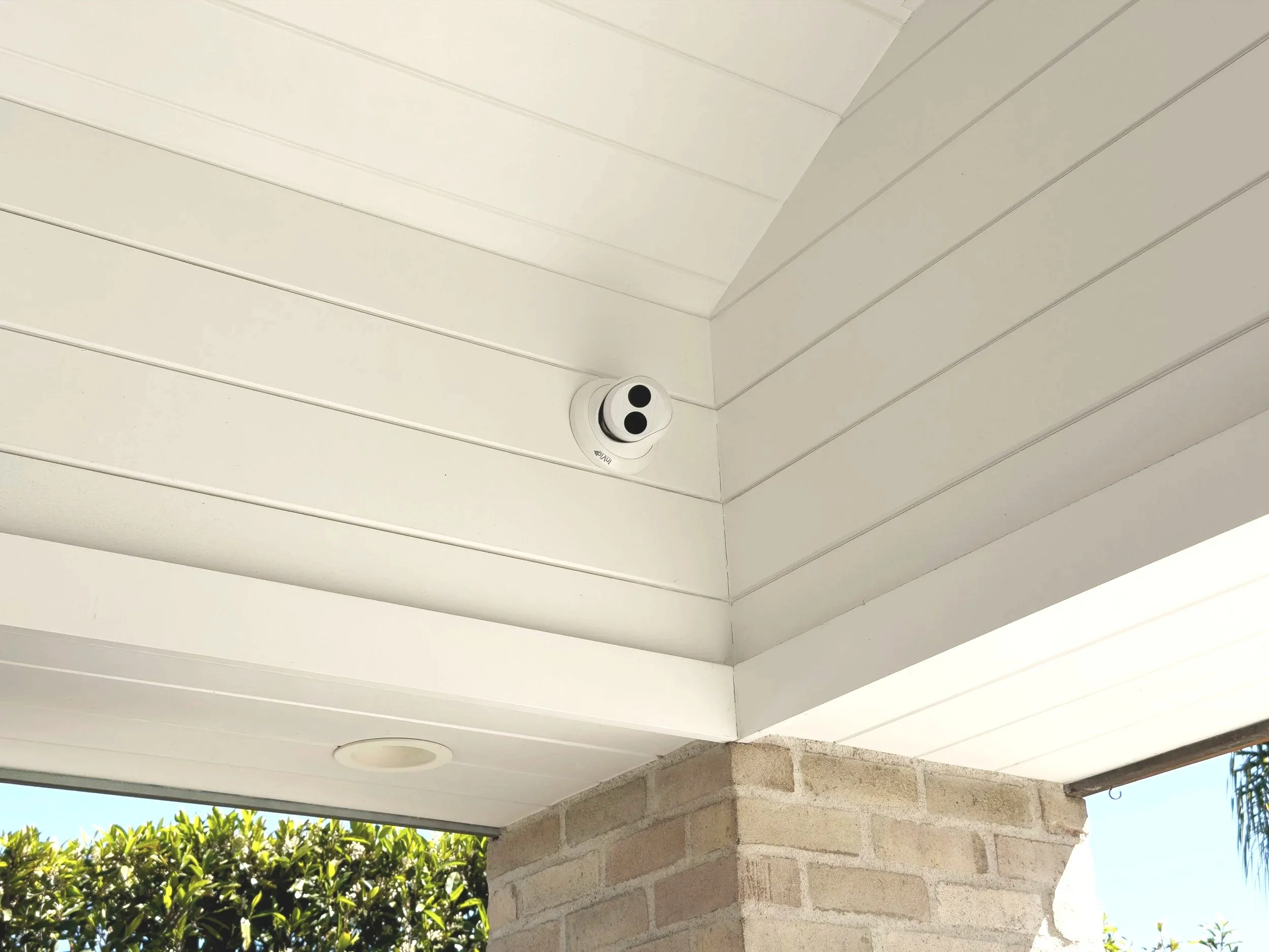 The image shows a white ceiling with a security camera mounted on it, a round recessed light, and a partial view of an exterior brick wall with green bushes and trees in the background.