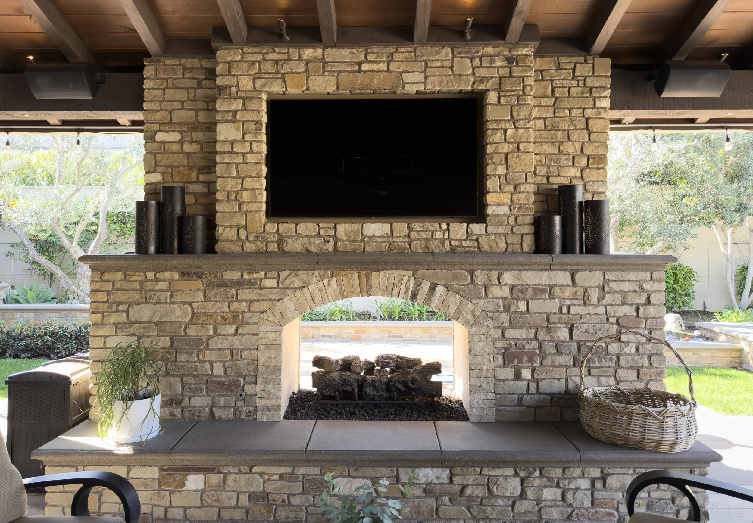 Outdoor stone fireplace with mounted TV and surround sound speakers by Surf City Audio Visual, patio entertainment setup