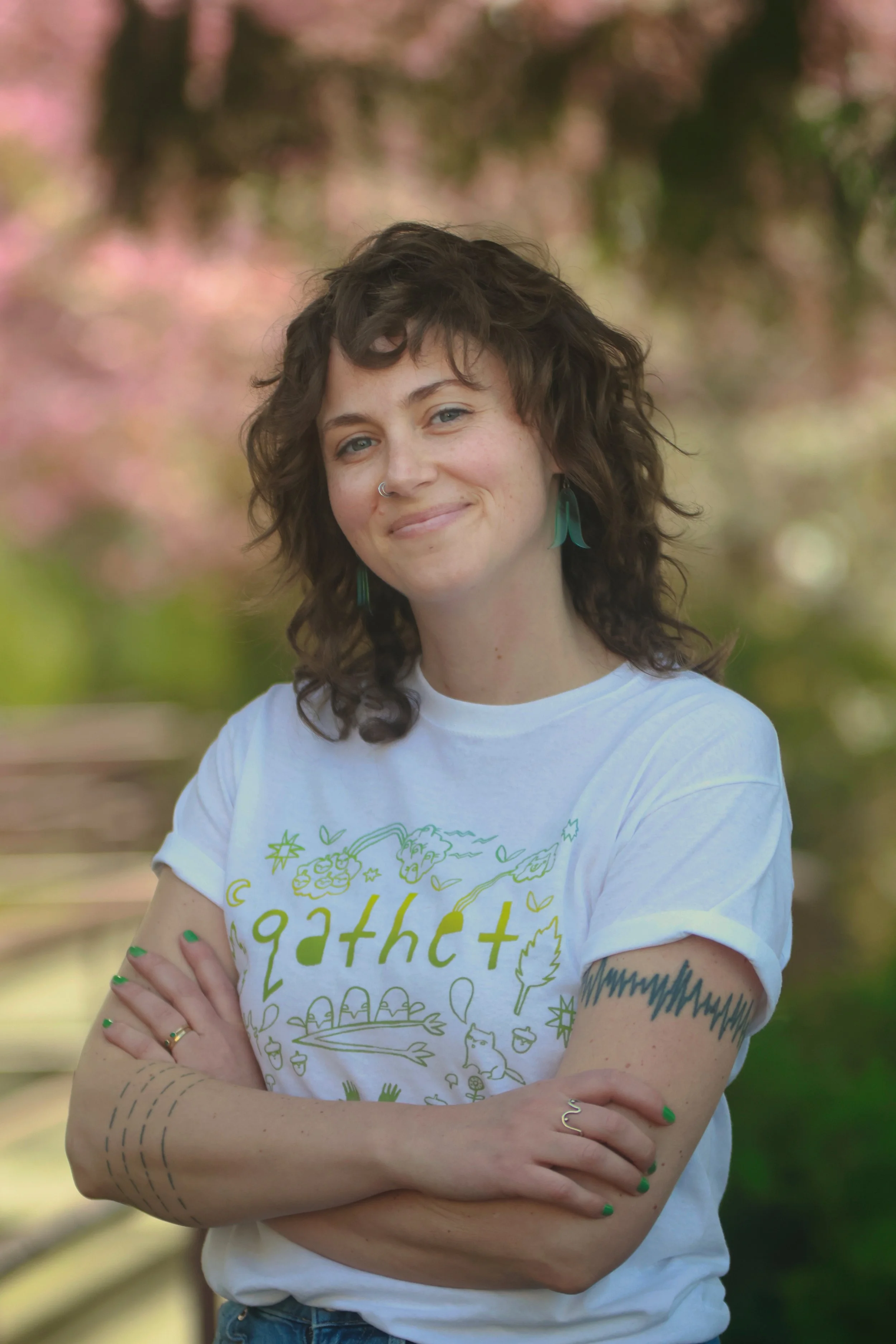A woman with curly brown hair, blue eyes, and tattoos standing outdoors with her arms crossed. She is wearing a white T-shirt with green illustrations and text, and turquoise earrings. The background is blurred with pink and green foliage.