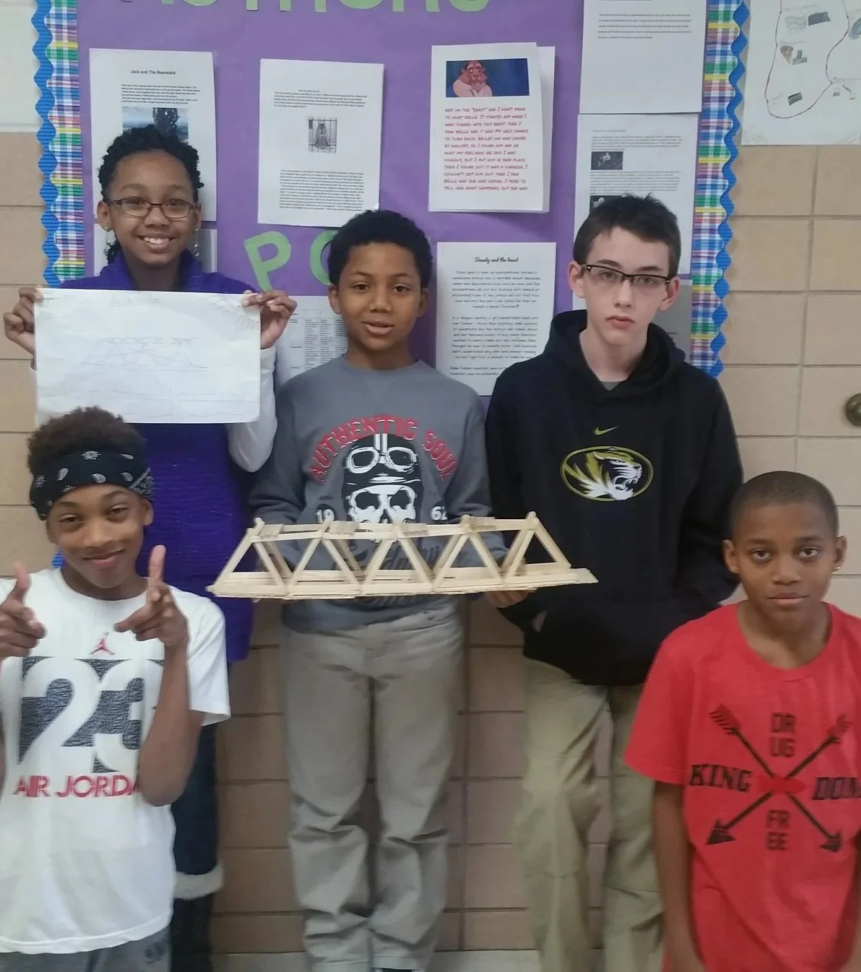 Group of five children standing in front of a purple bulletin board, holding a wooden bridge model, with some students smiling and posing for the photo.