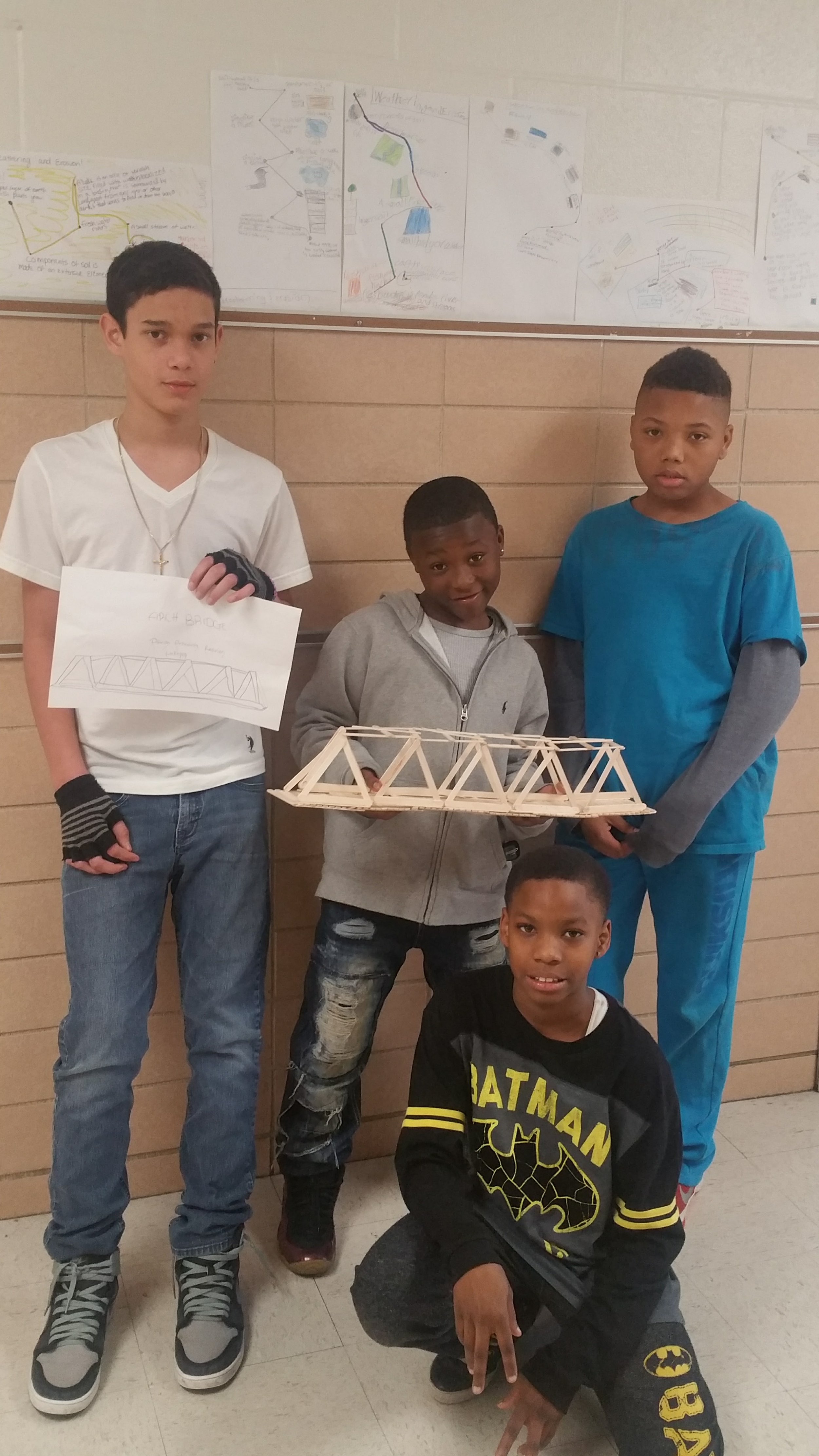 Five children standing in front of a beige wall with posters. One boy in a white shirt holds a paper with a sketch of a bridge. Another boy in a gray hoodie holds a small model of a bridge. Two other children, one in a blue shirt and another in a Batman-themed shirt, are nearby.