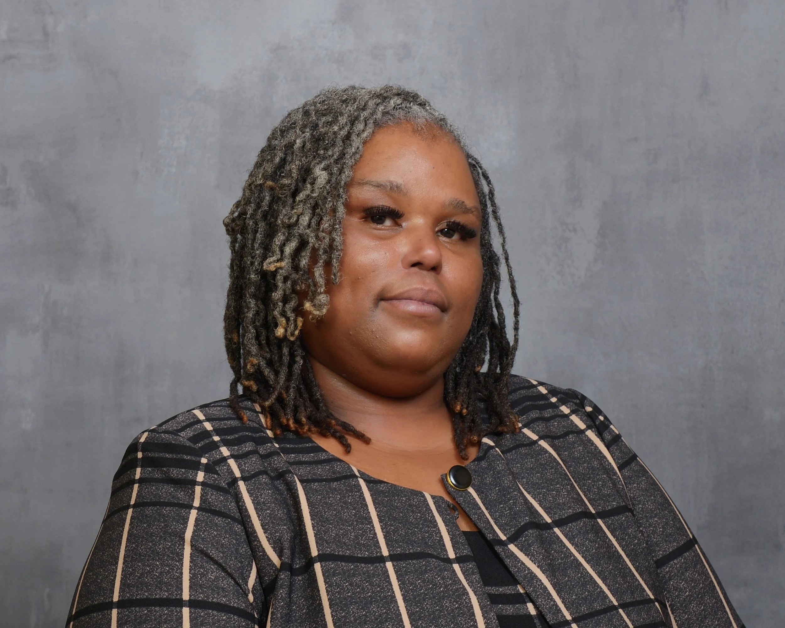 Close-up portrait of an African American woman with gray, textured dreadlocks, wearing a black and beige checkered top, standing against a gray background.
