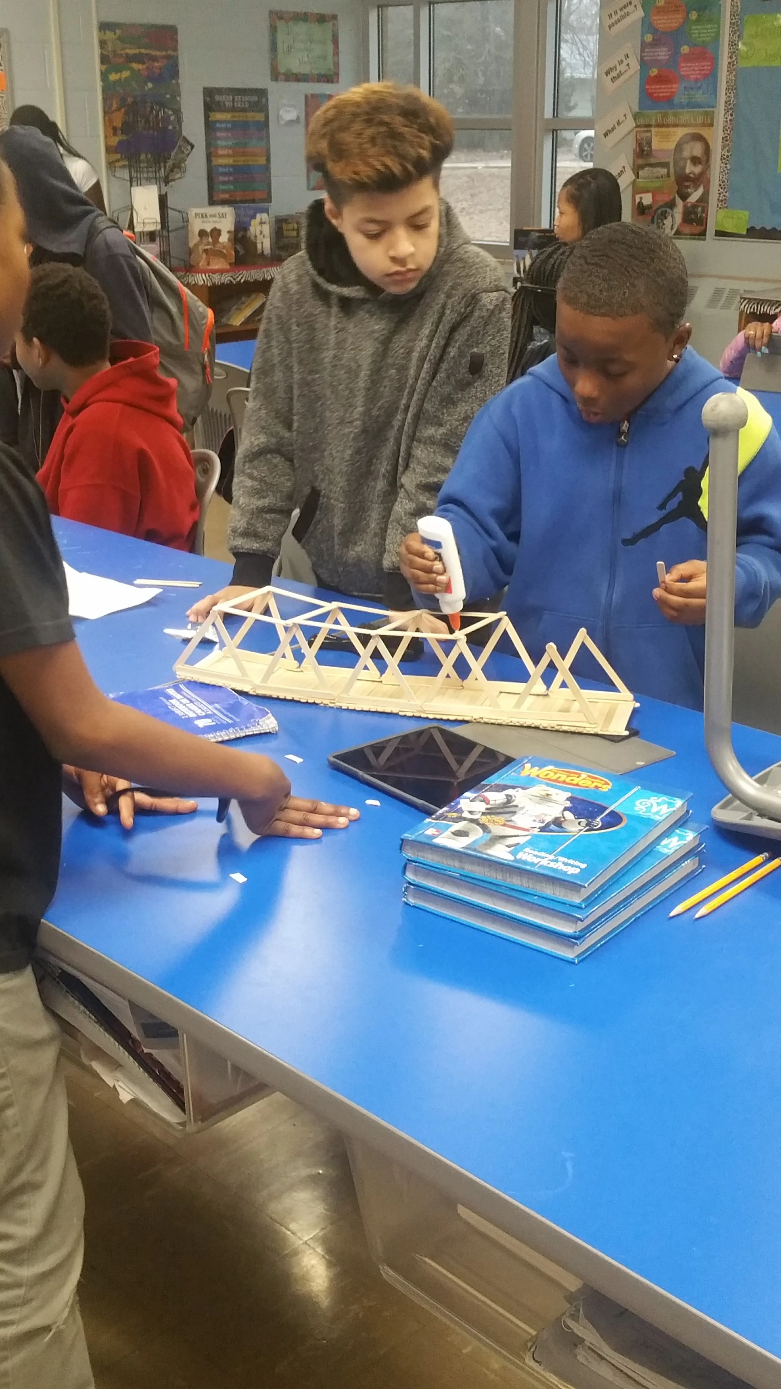 Two boys working on a wooden model bridge at a classroom table, surrounded by school supplies and books.