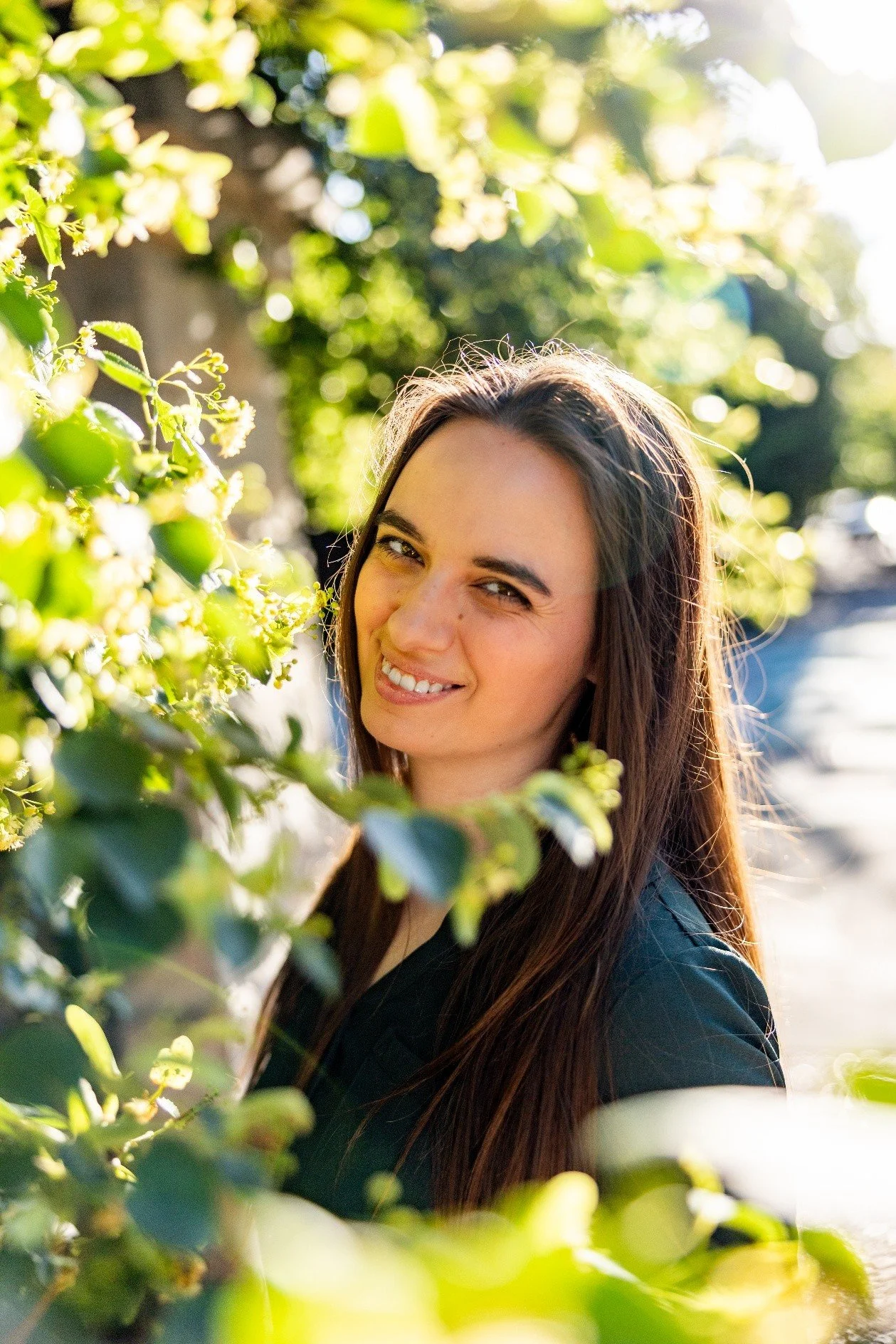 A young woman with long brown hair smiling outdoors in sunlight, surrounded by green foliage.