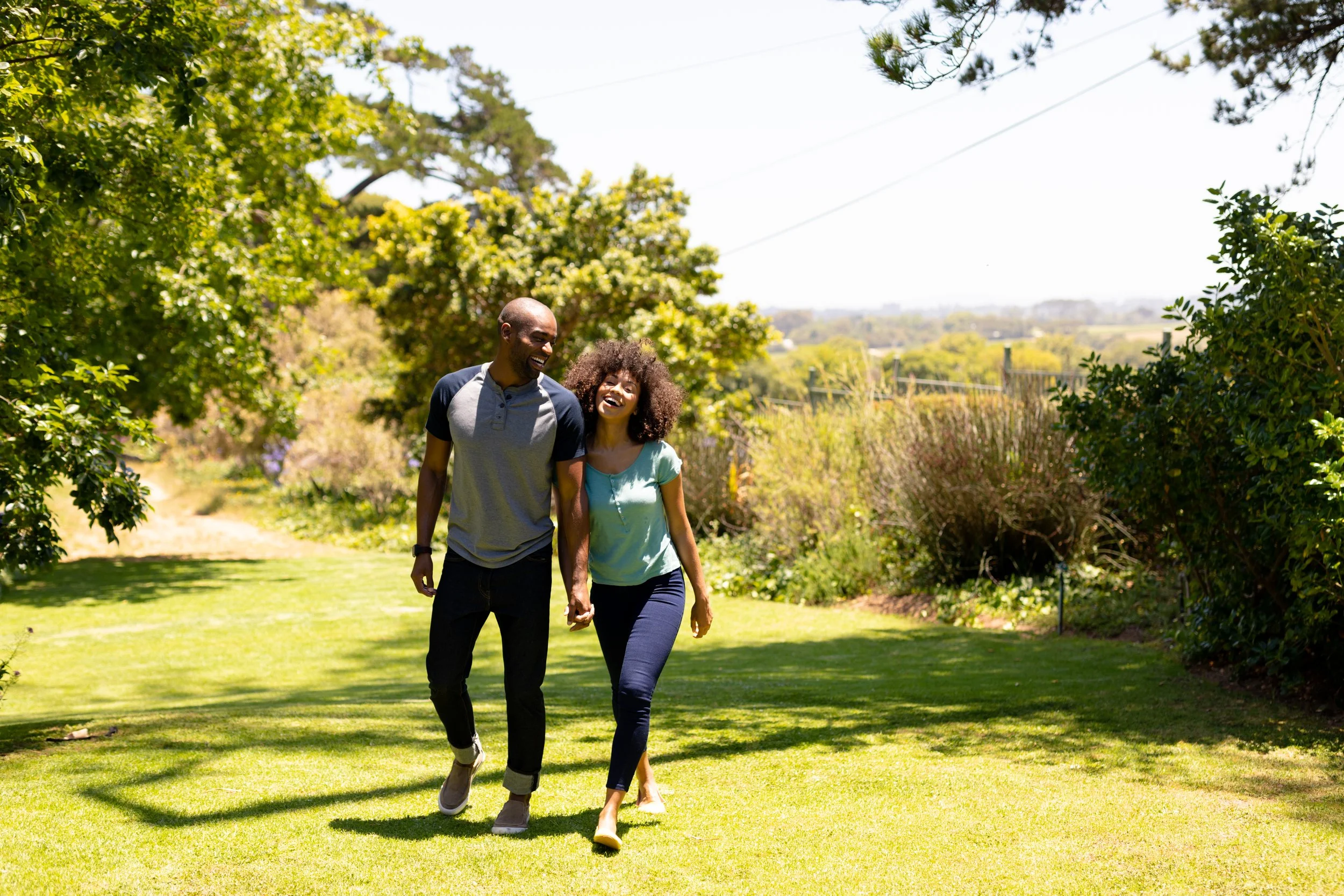 A man and a woman walking hand in hand on a sunny day in a grassy park surrounded by trees, smiling and enjoying each other's company.