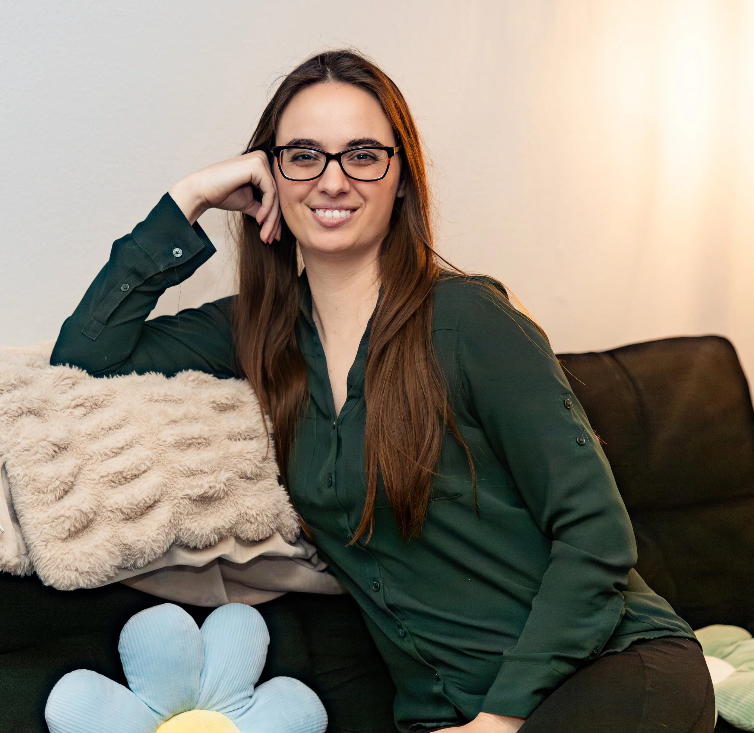 A woman with long brown hair, wearing glasses and a dark green shirt, is smiling and resting her head on her hand. She is sitting on a black sofa with a pink fuzzy blanket in front of her. There are several pastel-colored candles nearby, including yellow, green, and blue.