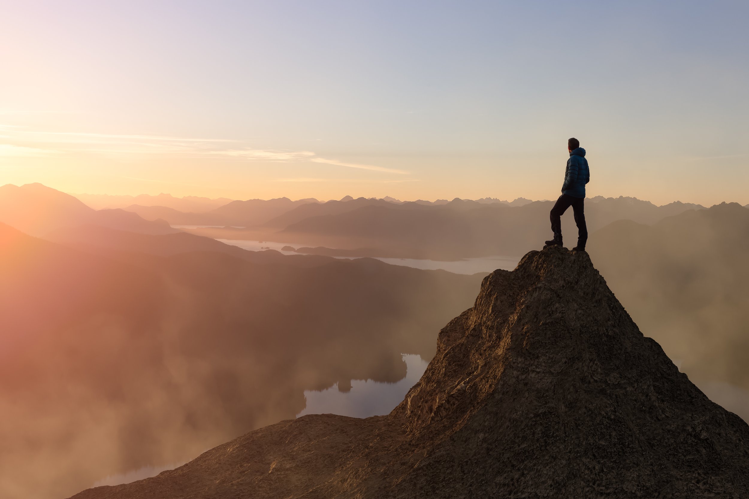 A person standing on a rocky ledge overlooking a mountain range and a body of water at sunset, wearing a blue jacket and black pants.
