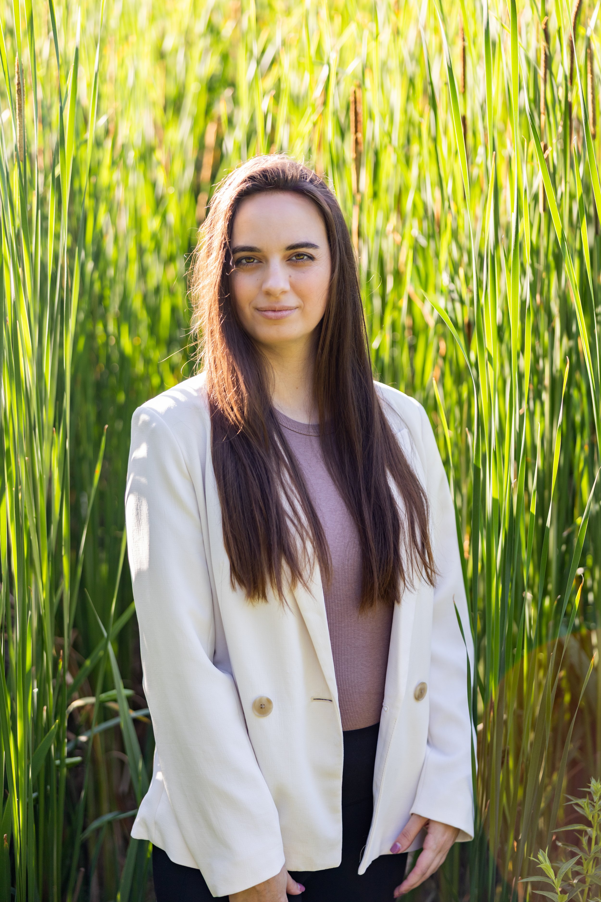A woman with long brown hair standing in a green field of tall grass, wearing a beige blazer and a purple top.