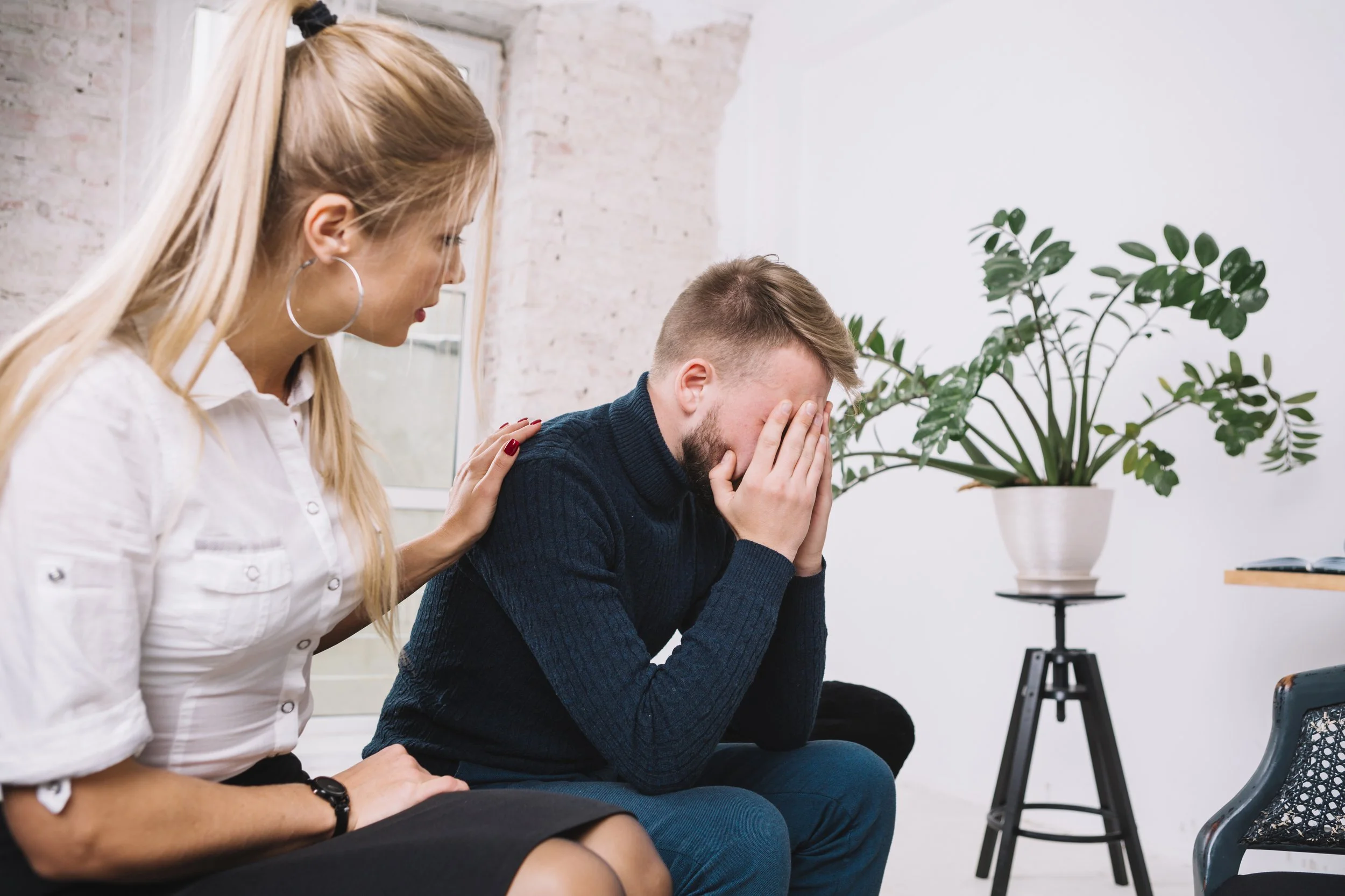A woman is comforting a man who is sitting with his face in his hands, appearing upset or distressed. The woman has long blonde hair, large hoop earrings, and a white shirt. The scene is in a bright room with a large potted plant in the background.