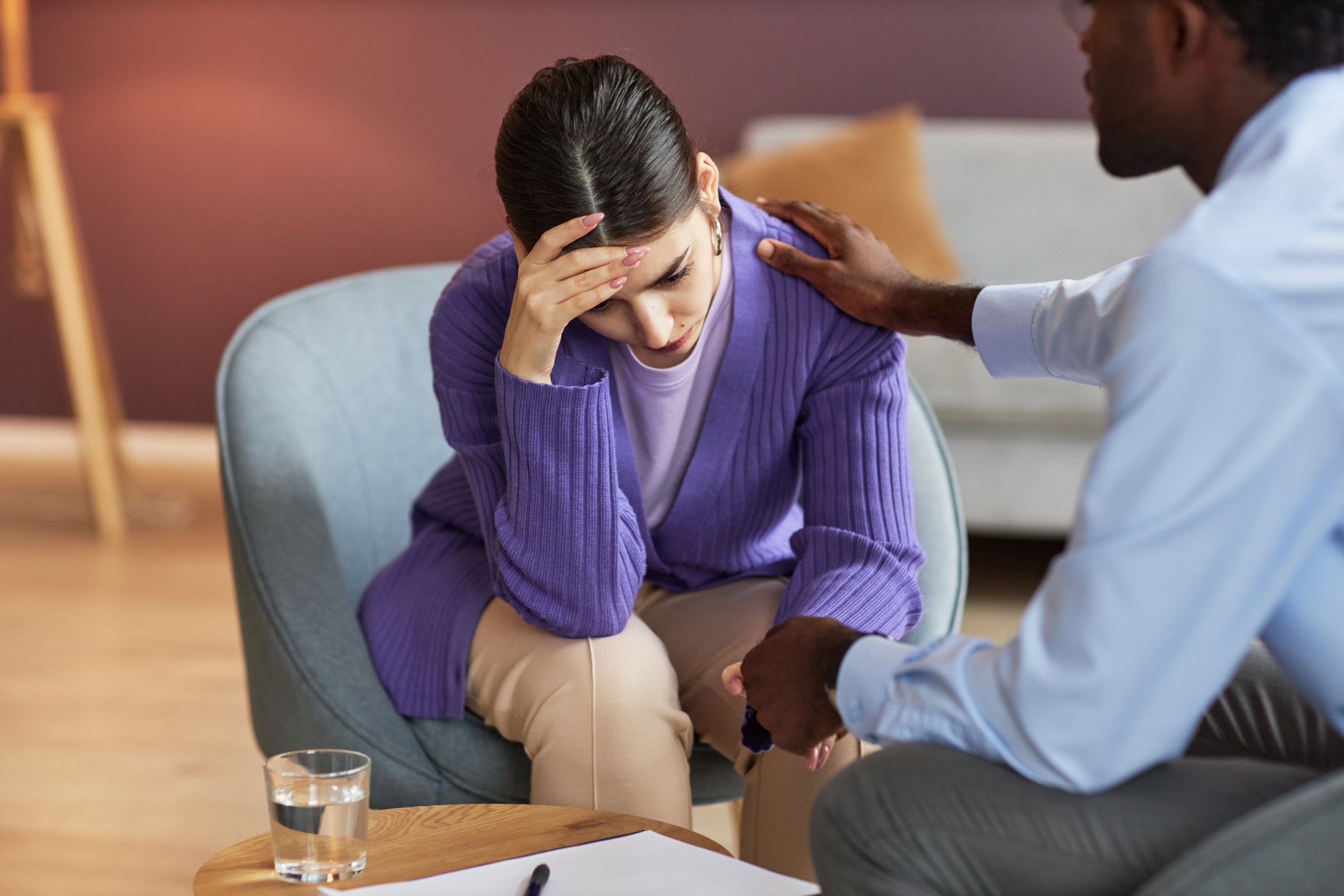 A woman sits with her head in her hand, appearing distressed, while a man, possibly a doctor or counselor, comforts her by placing a hand on her shoulder. They are seated in a room with warm lighting, and a glass of water, paper, and pen are on a table in front of them.