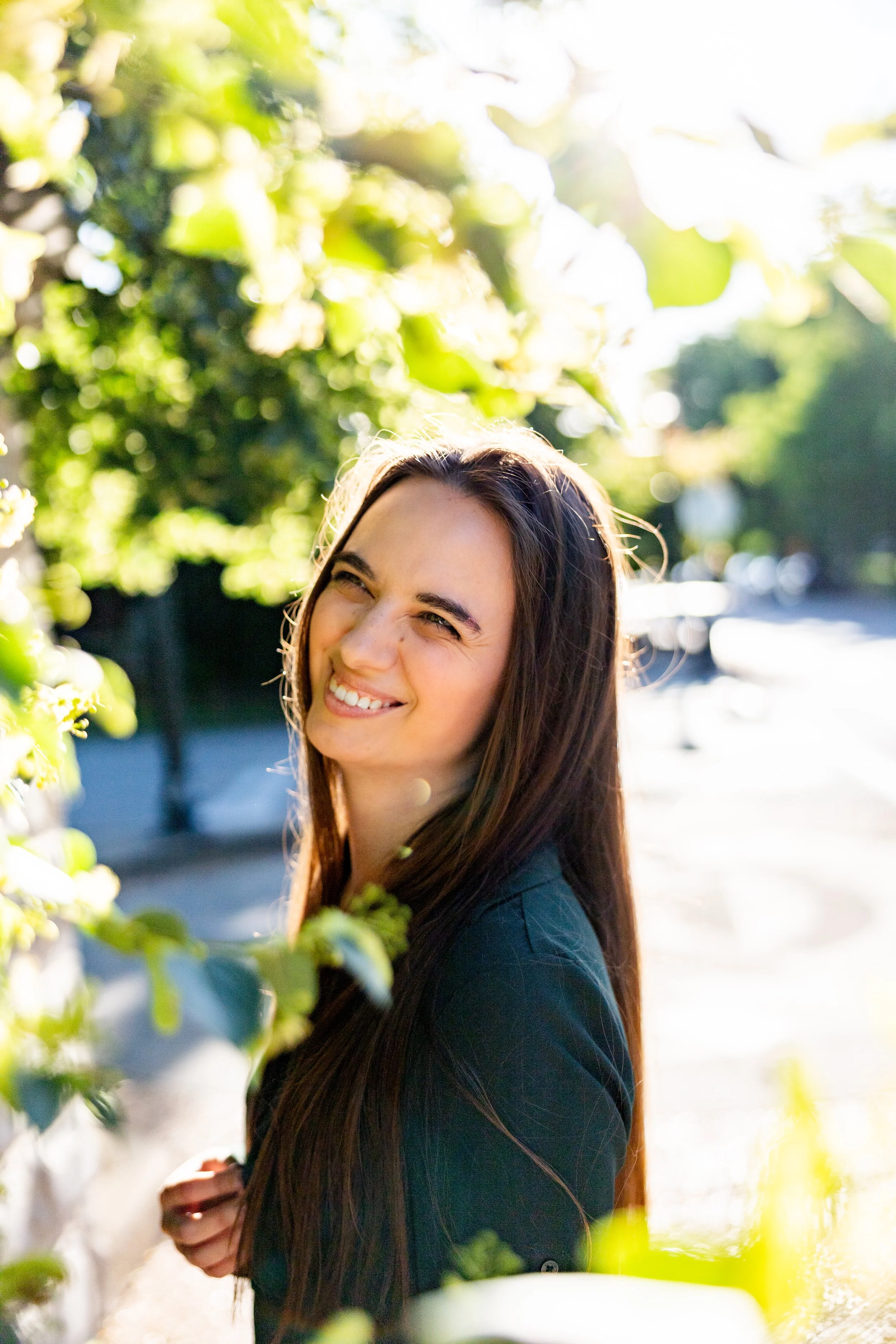 A young woman with long brown hair smiling outdoors among greenery, with sunlight shining through leaves in the background.