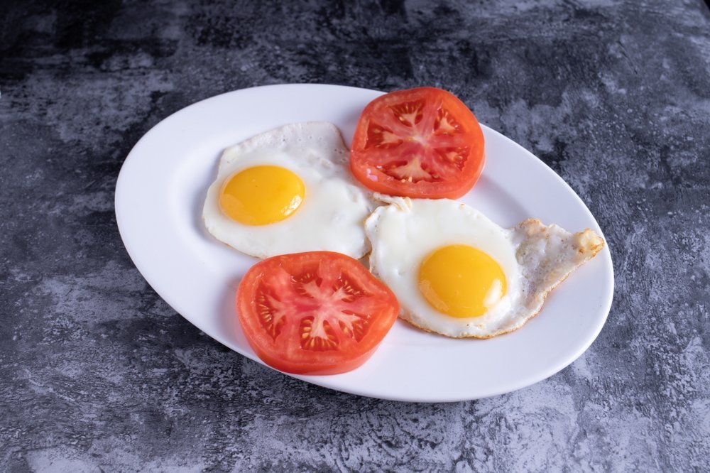 Two fried eggs and two slices of tomato on a white plate, placed on a dark gray surface.