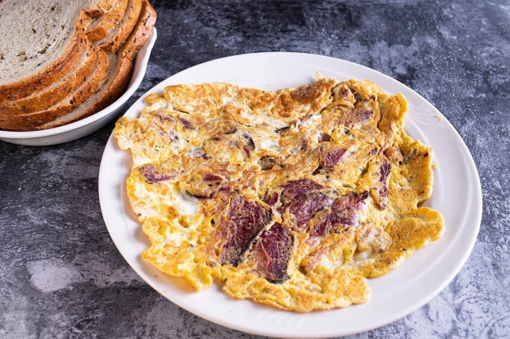 Fried egg with bacon on a white plate and slices of bread in a bowl on a dark gray surface.