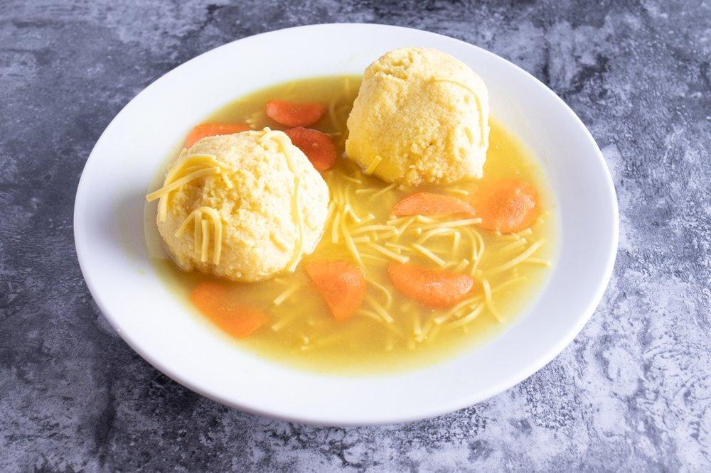 A bowl of chicken noodle soup with two large matzo balls, sliced carrots, and shredded cheese on top, on a dark gray textured surface.