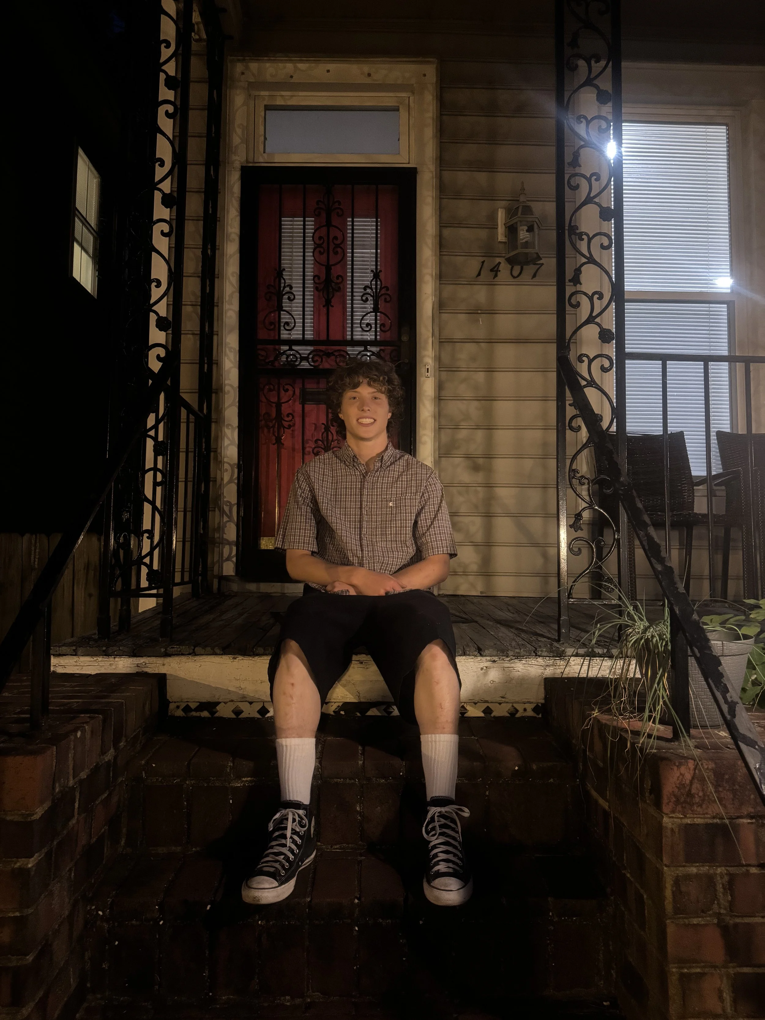 A young man with curly hair is sitting on the steps of a porch at night. His legs are visible, showing bruises and scrapes. He is smiling and wearing a brown checkered shirt, black shorts, white socks, and black and white sneakers. The porch has black metal railings and a red door with a black security grill, flanked by a window with blinds. The house number 1407 is visible on the wall next to a wall-mounted lantern.