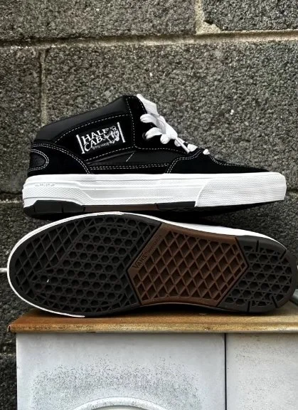 A pair of black, white, and brown sneakers with white laces, placed on a wooden surface against a brick wall. The sneakers display the brand name Halex.