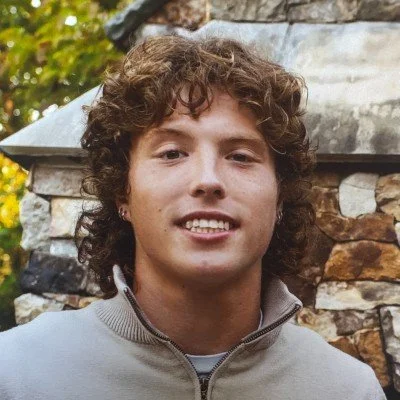 A young man with curly brown hair smiling outdoors in front of a stone wall and greenery.