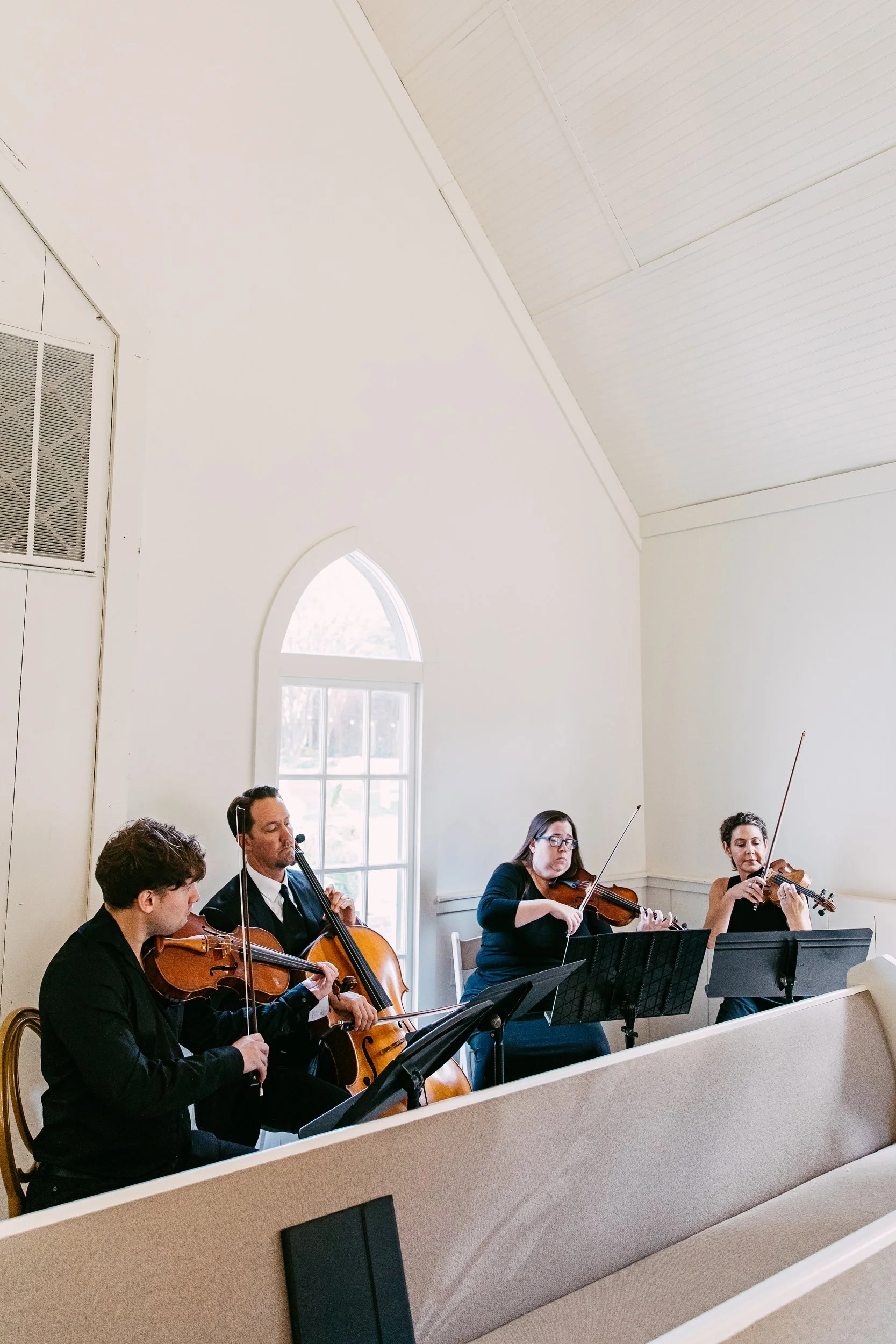 A group of musicians playing string instruments, including violins and cellos, during an indoor performance in a bright room with large windows and high, white ceiling.