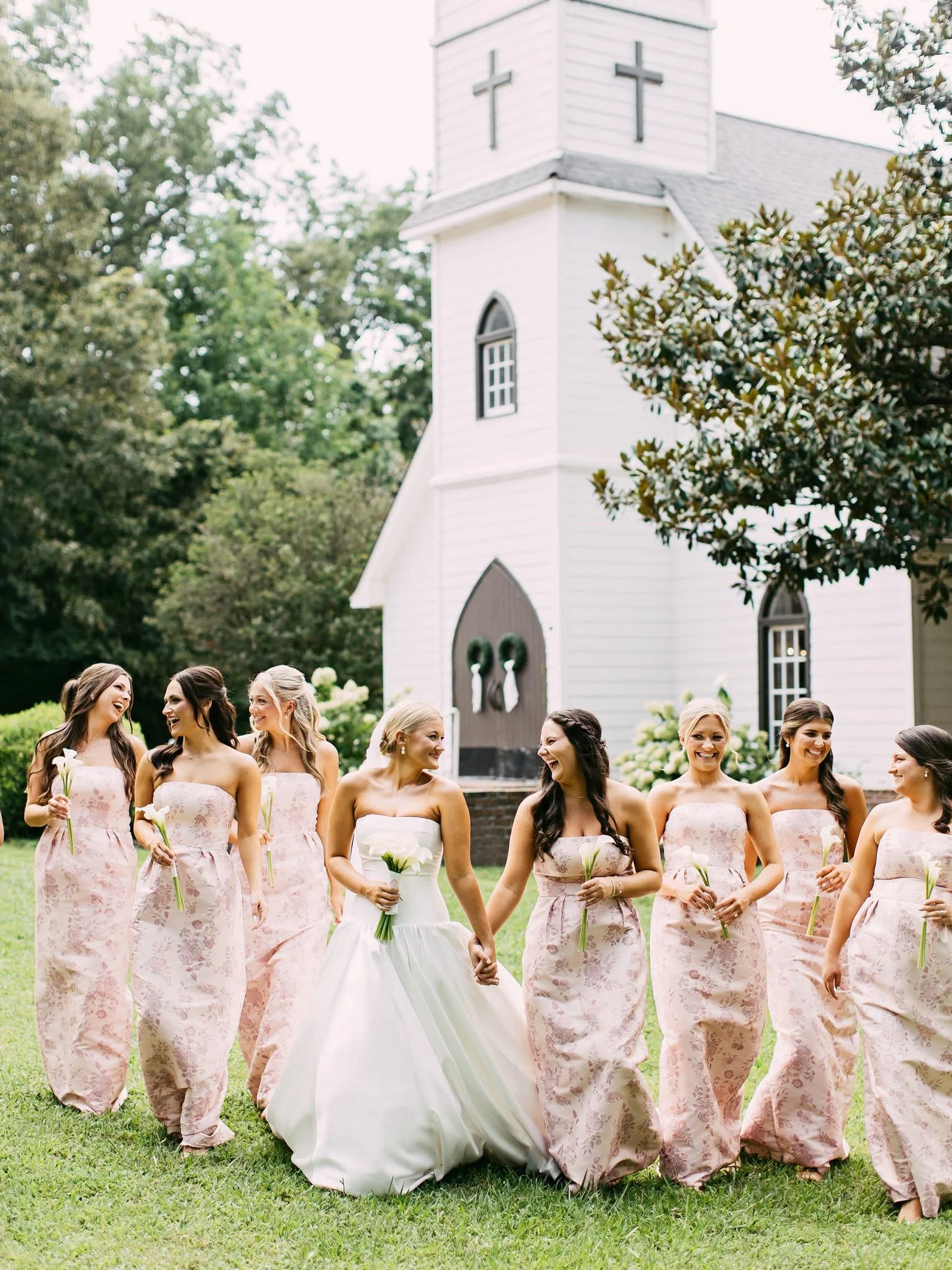 A bride in a white wedding dress holding hands with her bridesmaids in pink floral dresses. The bridesmaids are holding white calla lilies. They are standing on a green lawn in front of a white church with a steeple.