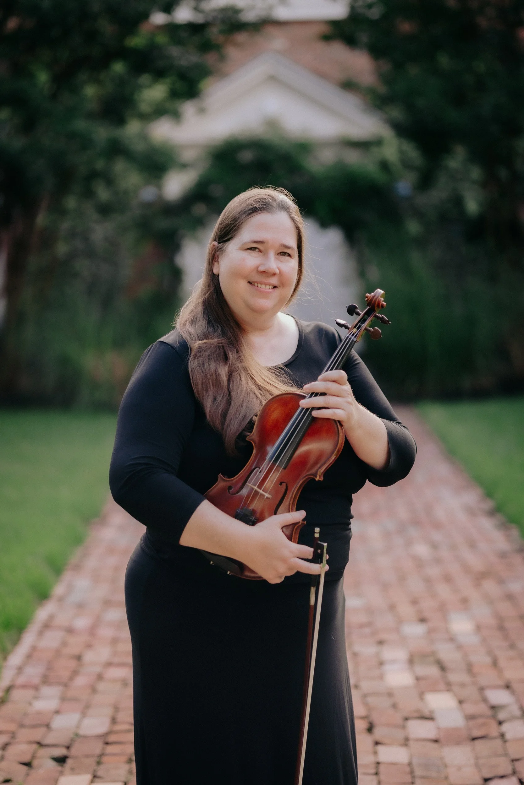 A woman with long brown hair smiling while holding a violin and bow, standing on a brick pathway outdoors with trees and a white building in the background.