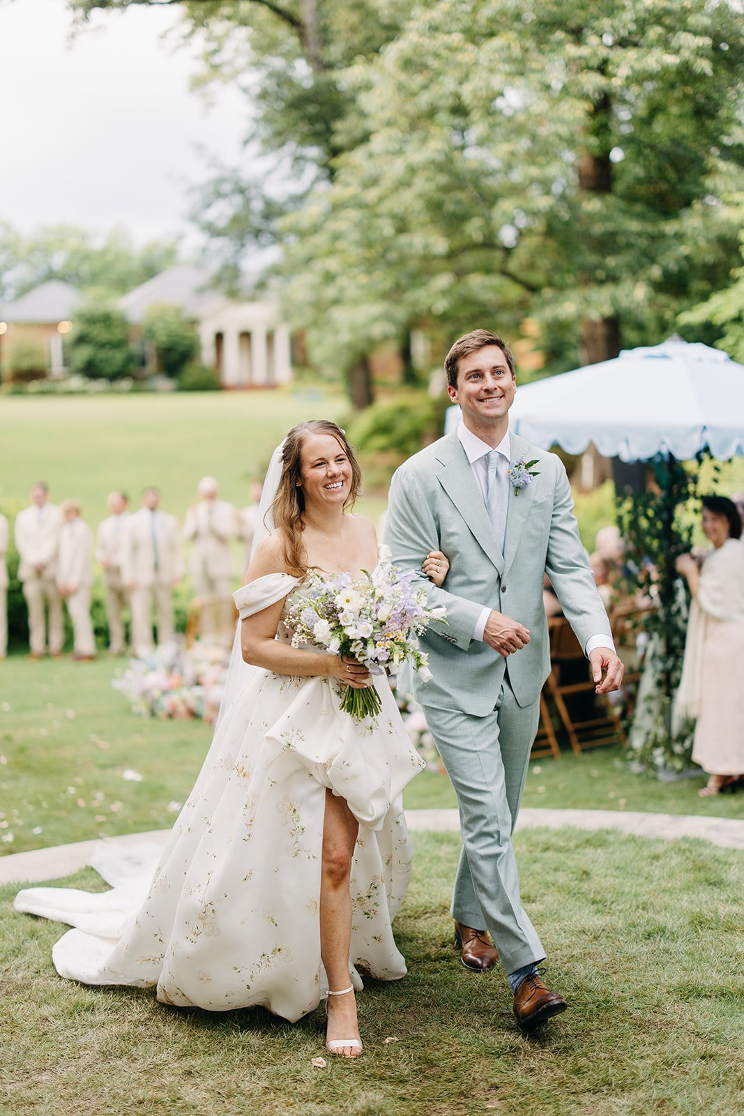 A bride and groom walk down the aisle during an outdoor wedding ceremony with guests and a tent in the background.
