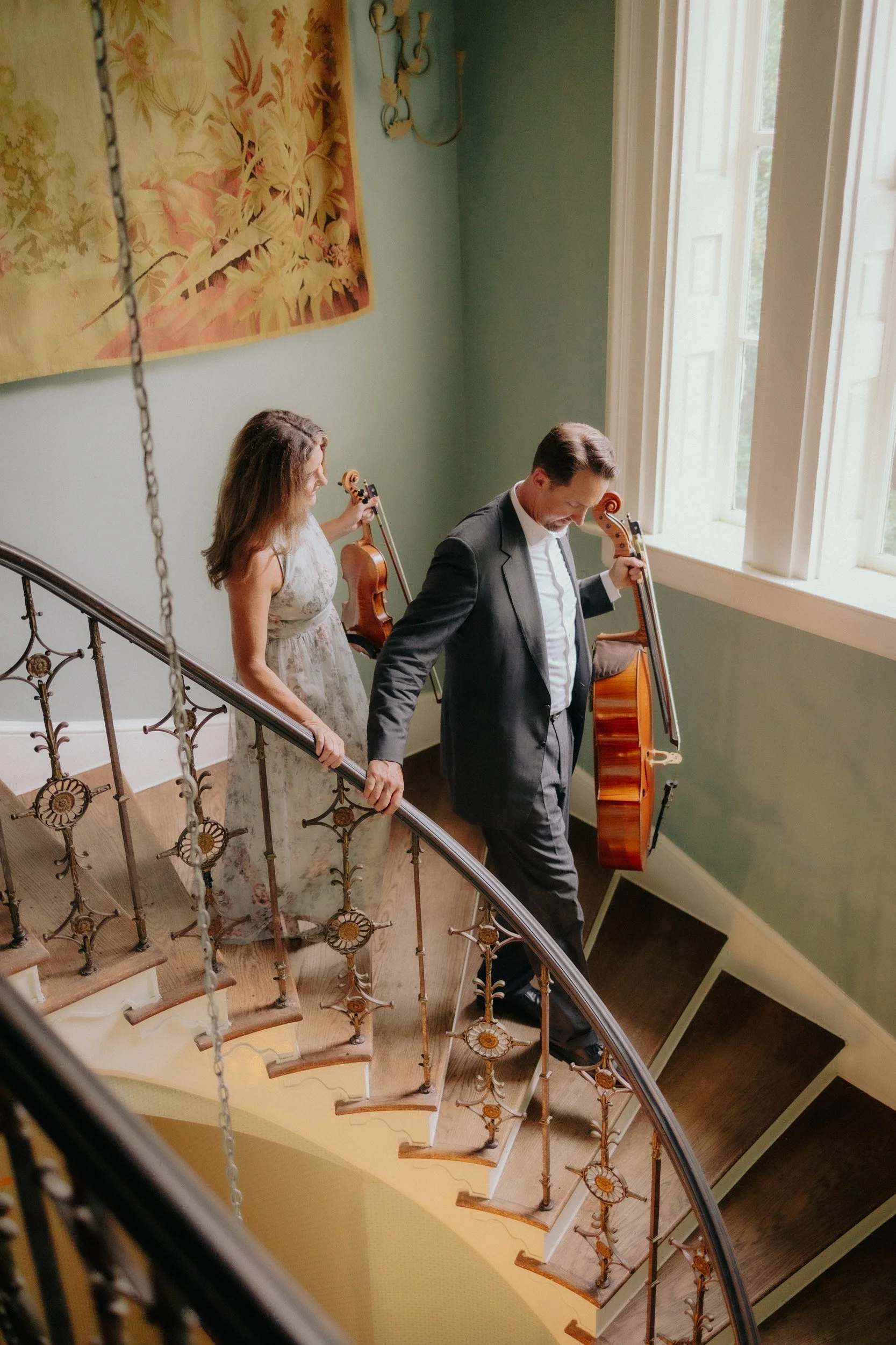 A man in a suit and a woman in a floral dress standing on a staircase with a violin and a cello, near an open window with sunlight streaming in, in a room with green walls and a large floral painting hanging on the wall.