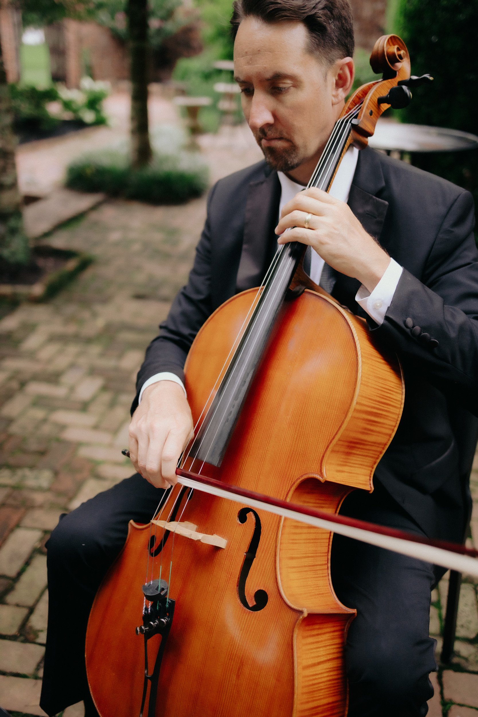 A man in a black suit playing a cello outdoors on a brick path surrounded by greenery.